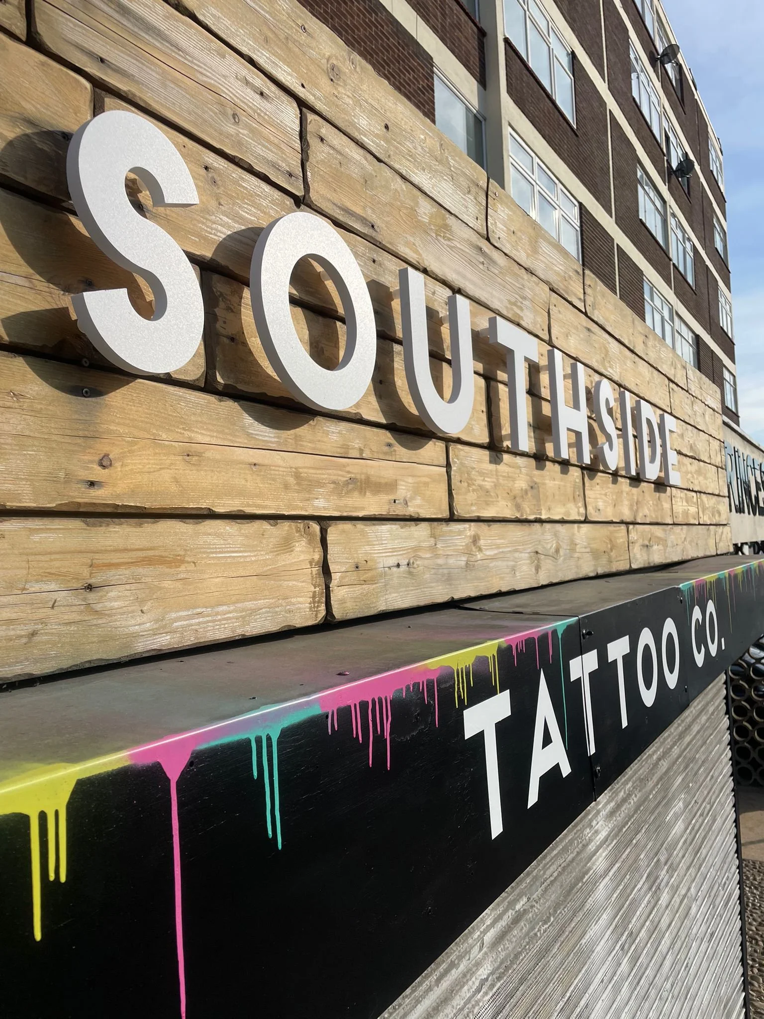 Close-up of a sign that reads 'SOUTHSIDE' mounted on a wooden panel with a business sign below that reads 'TATTOO CO.' with colorful paint drips.