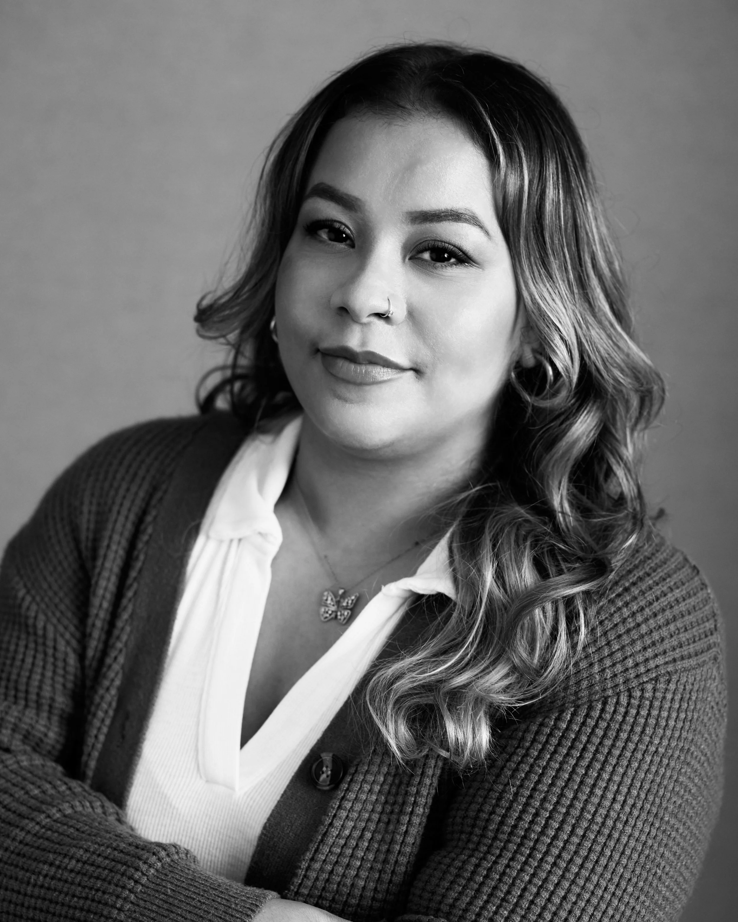 Black and white portrait of a woman with wavy hair wearing a white blouse, cardigan, and butterfly necklace. She has a nose ring and earrings.