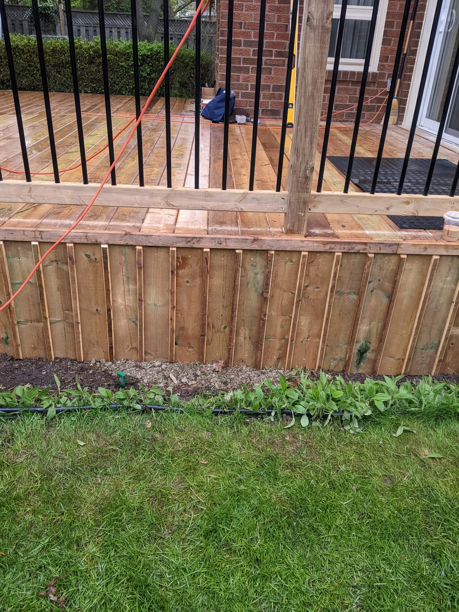 A new wooden deck under construction with black metal railing, a brick house in the background, and a grassy area with plants in the foreground.