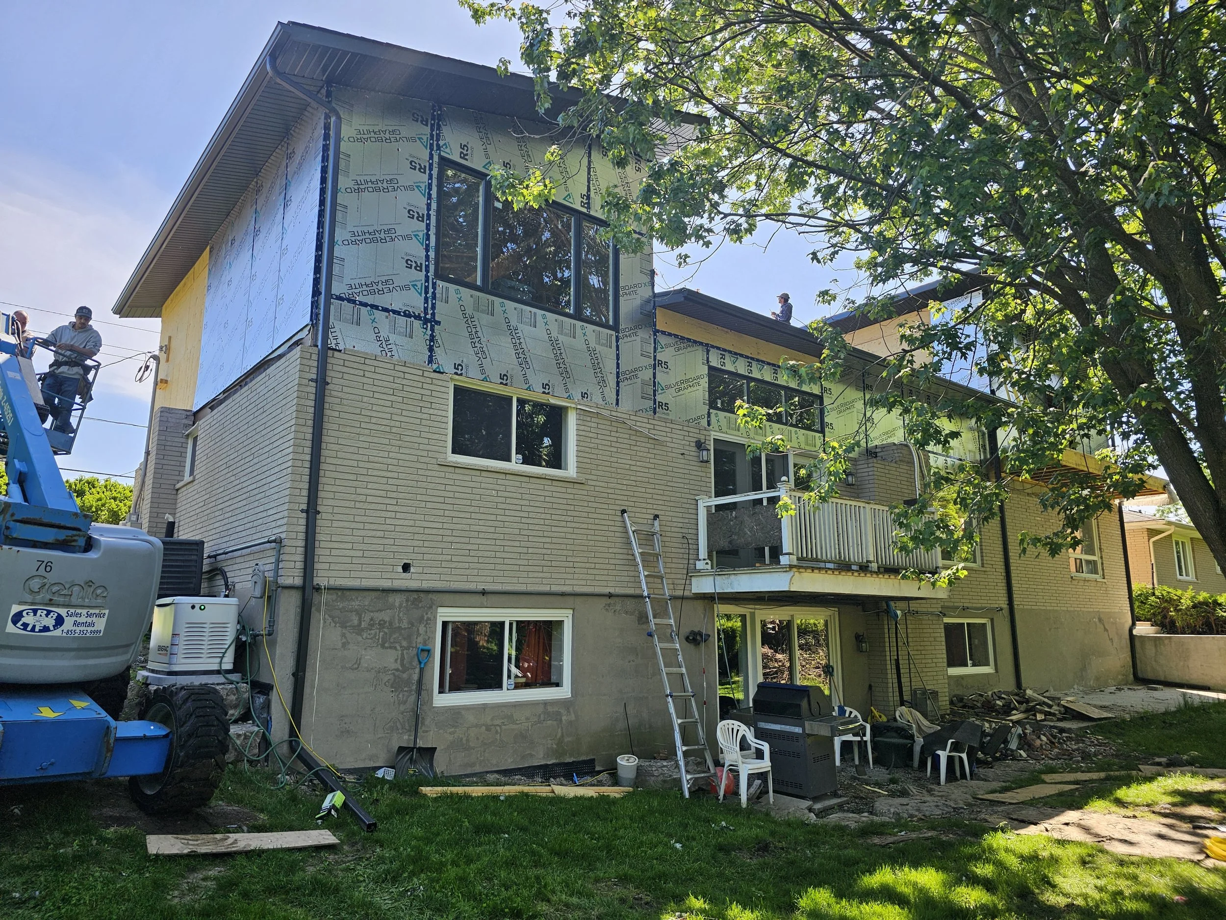 A multi-story house under construction with workers and equipment, partially covered with insulation panels, surrounded by trees and construction materials.
