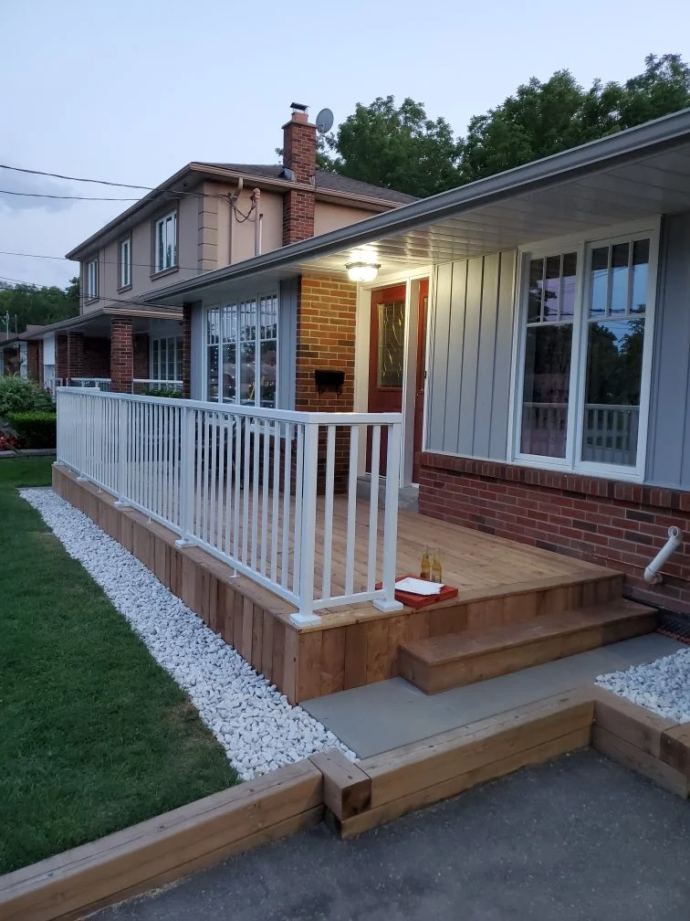 Exterior view of a house with a small porch, white railing, brick and siding facade, and a wooden deck. The porch has two steps leading down to a walkway, with white decorative stones along the edge. The house has large windows and a red door, with a