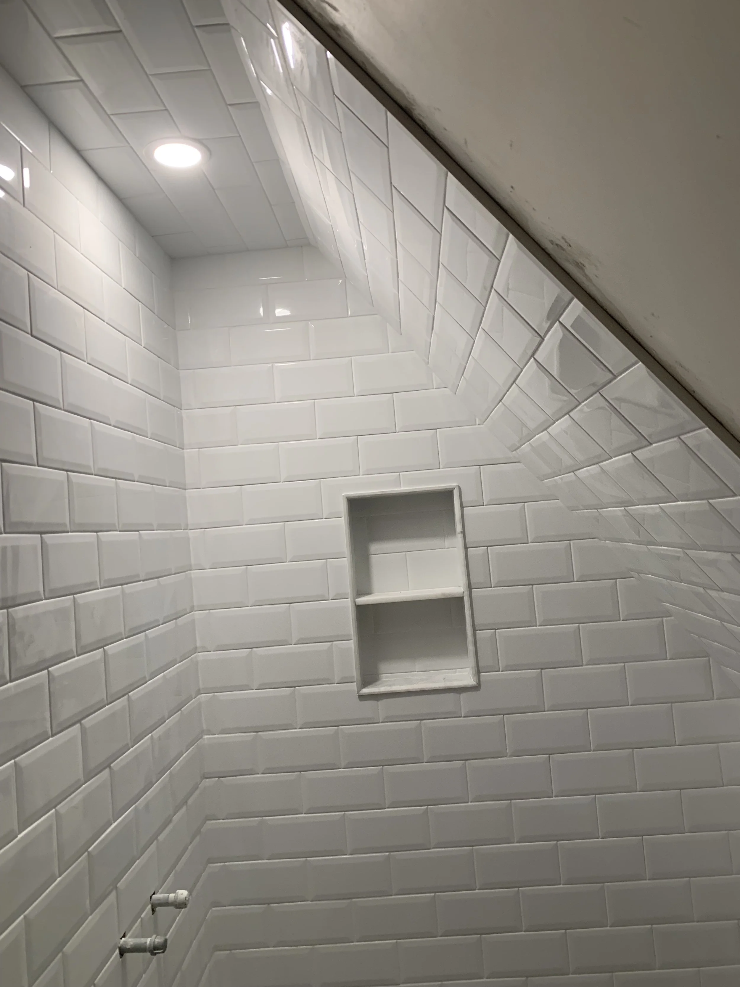 Bathroom with white subway tile walls, a small recessed shelf, a ceiling light, and part of the ceiling visible with some dirt or mold.