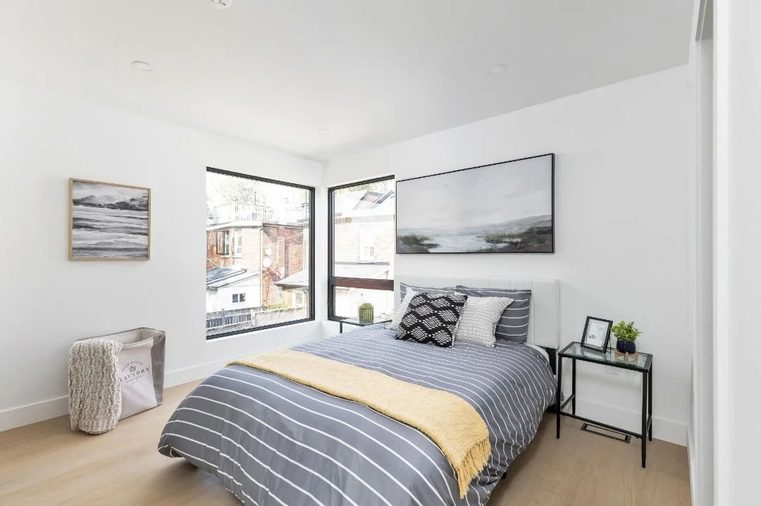 A modern bedroom with white walls, a large window, a bed with gray and white striped bedding, black and white pillows, a yellow blanket, artwork on the walls, a small side table with a potted plant, frame, and decorative item, and a laundry basket.