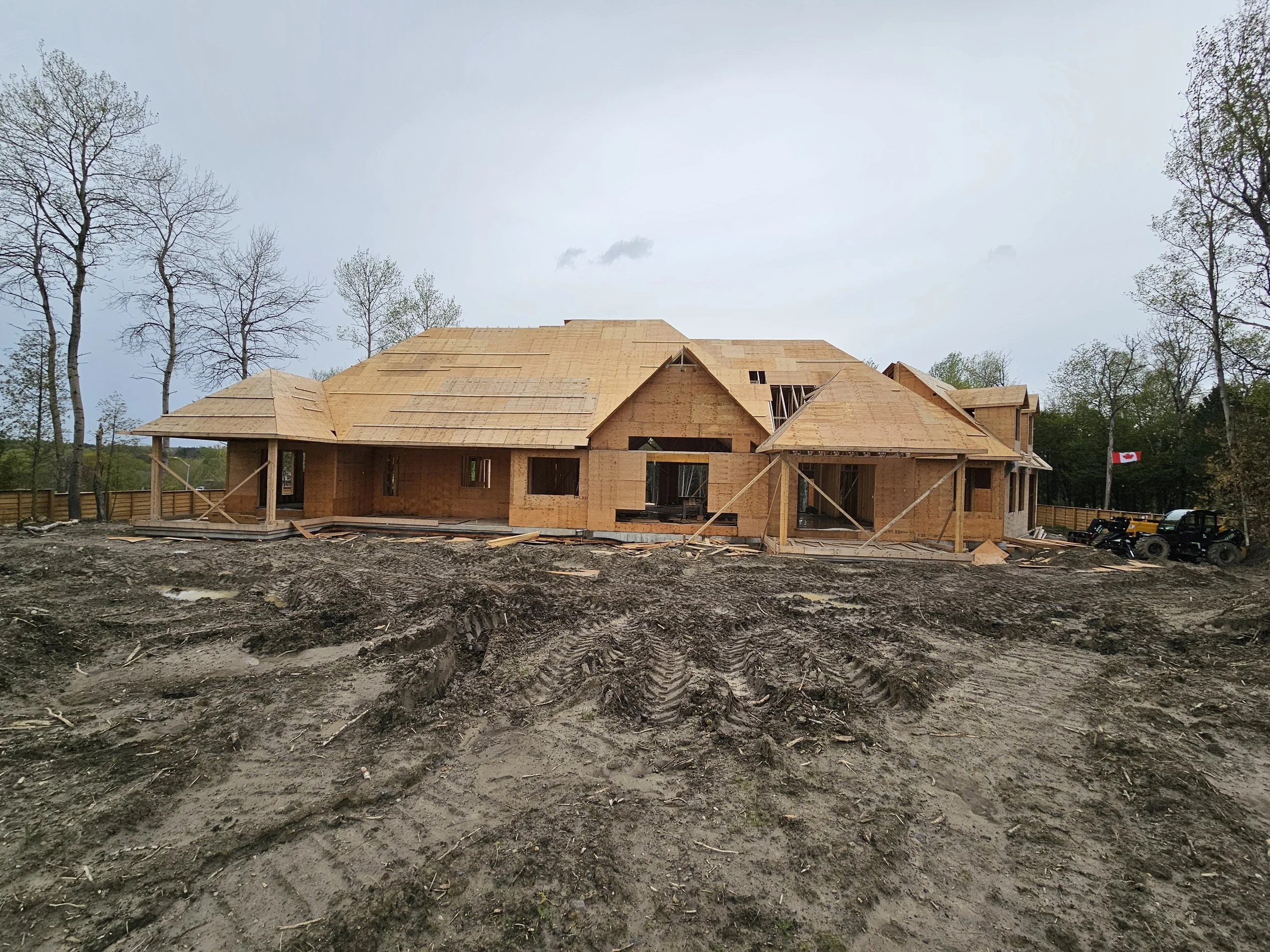 A house under construction with wooden framing and roof trusses, surrounded by dirt and construction equipment, with trees and a cloudy sky in the background.