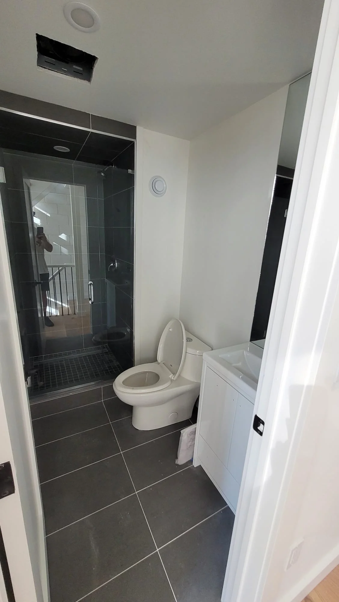 A bathroom with a glass-enclosed shower, a white toilet, a small white sink cabinet, and a black and white tile floor.