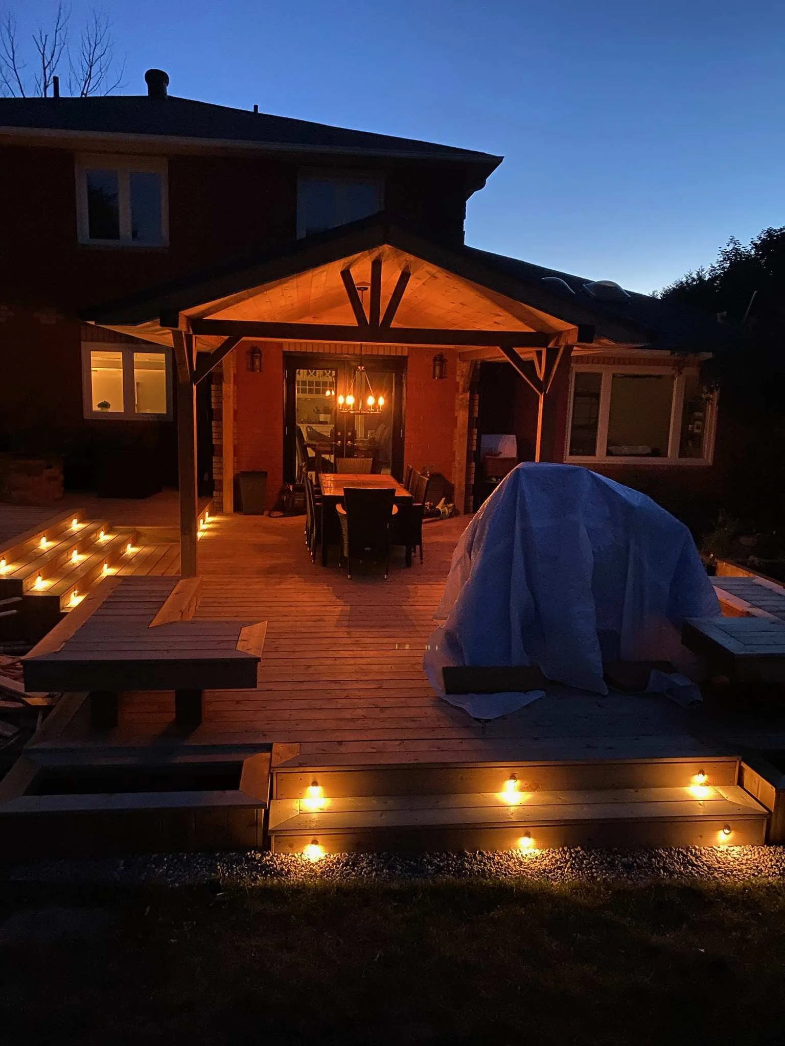A lit outdoor wooden deck with steps and string lights, attached to a house with large windows, during dusk or night.