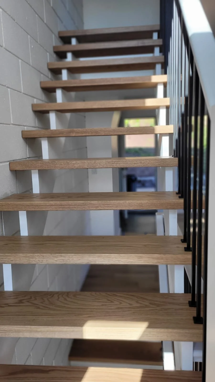 Interior view of a staircase with wooden steps, white support beams, black handrails, and a brick wall on the left side.