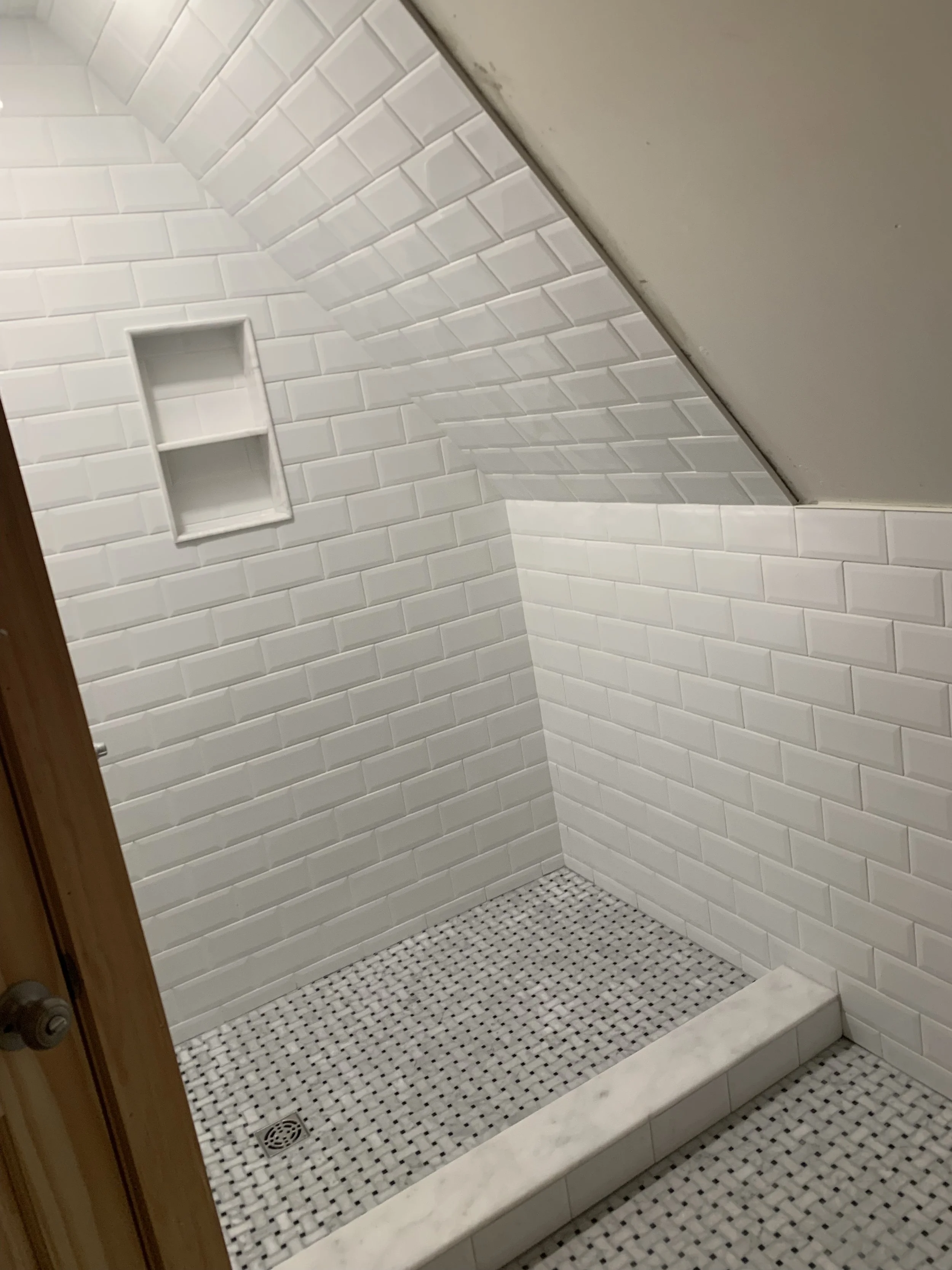 A small shower with white subway tiles on the walls, a built-in niche, a sloped ceiling, and a black-and-white mosaic tile floor.