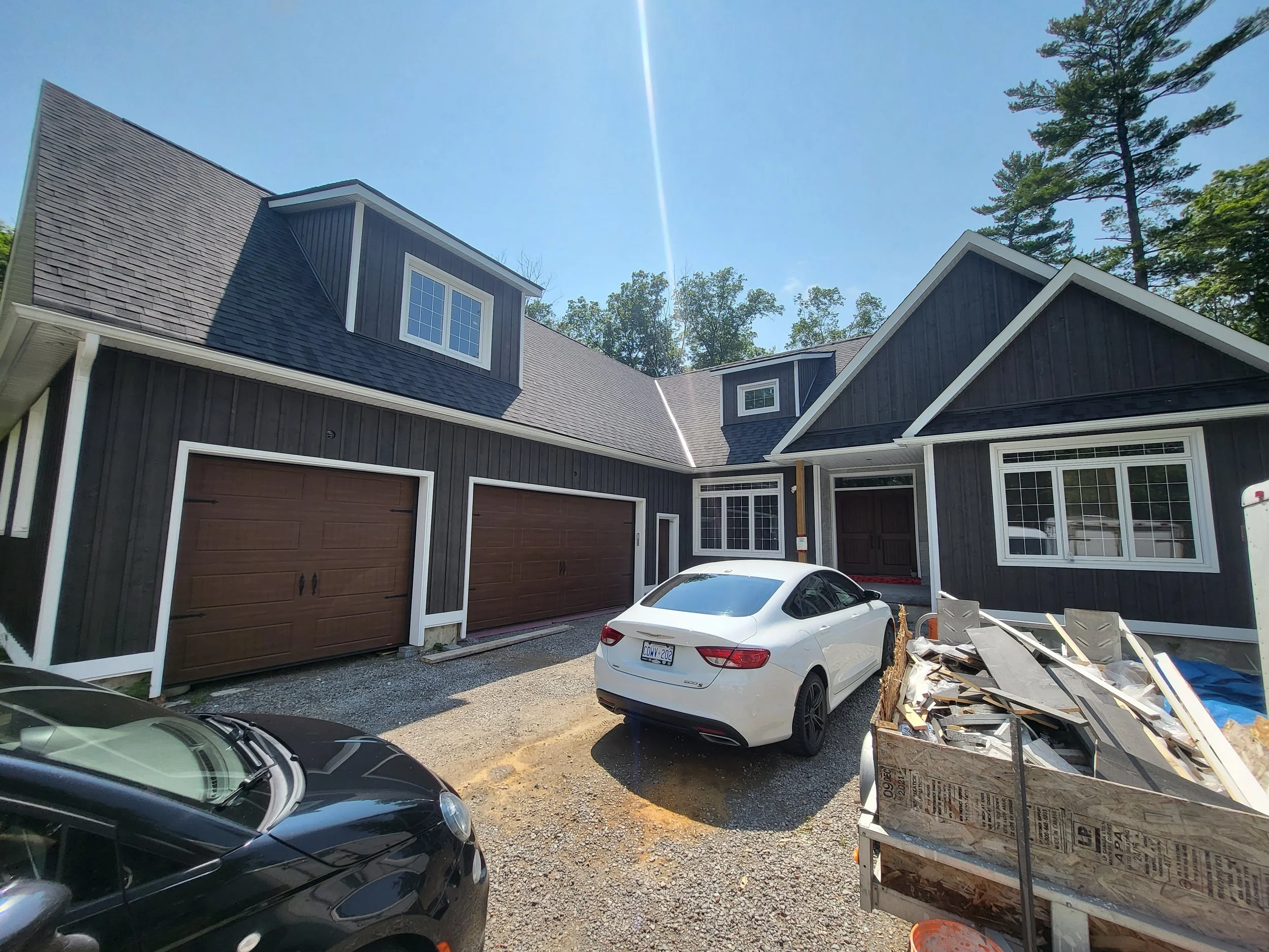 New two-story black house with attached garage and cars parked in front, surrounded by construction debris on a sunny day with clear blue skies.