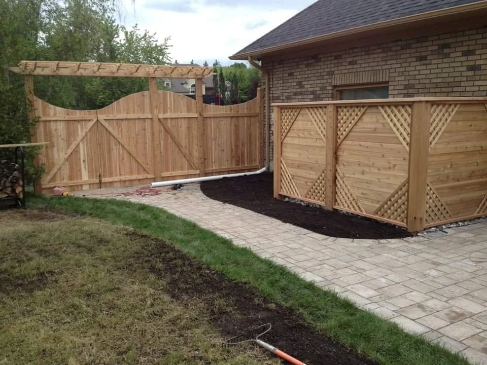 A backyard with a newly installed wooden fence, a paved walkway, and a brick house. The fence has lattice accents and a gate, with some gardening work in progress.