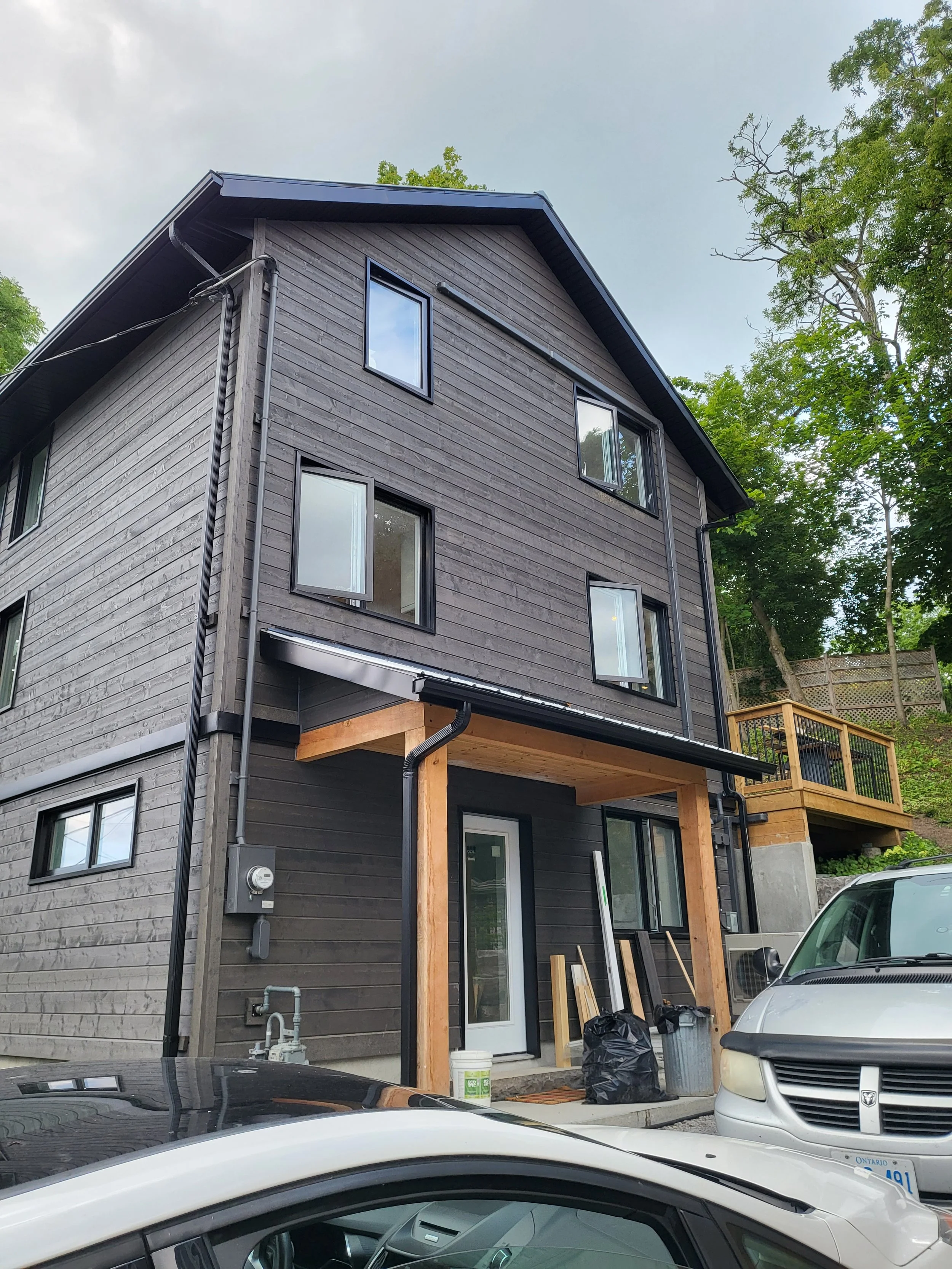 Newly constructed multi-story house with dark wood siding, multiple windows, a small covered porch, and a wooden deck on the side, surrounded by trees and parked cars.