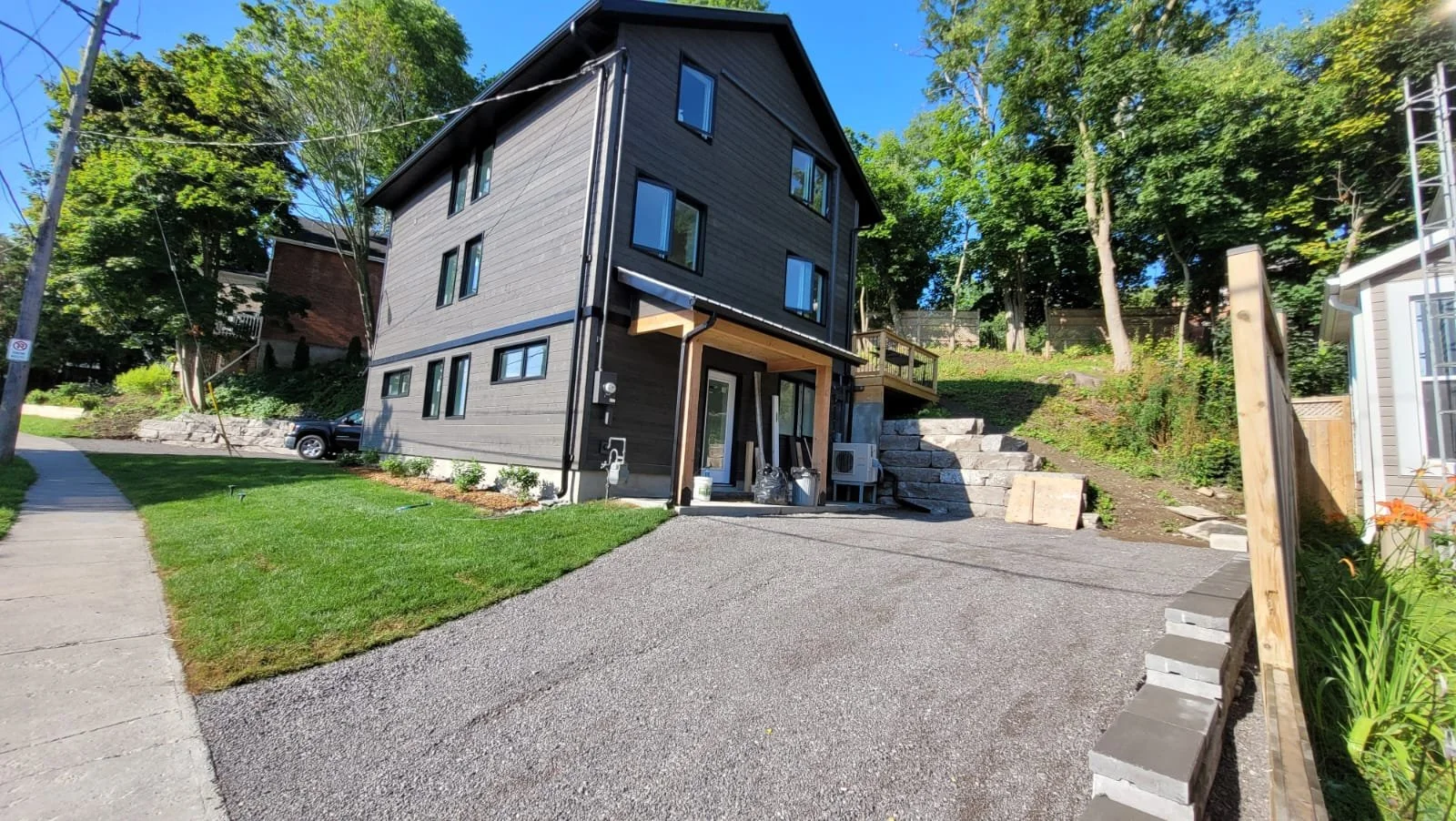 A modern, three-story house with a dark gray exterior, multiple windows, a small front yard with green grass, a concrete sidewalk, and a gravel driveway. The house is situated on a sloped lot with trees and greenery surrounding it.
