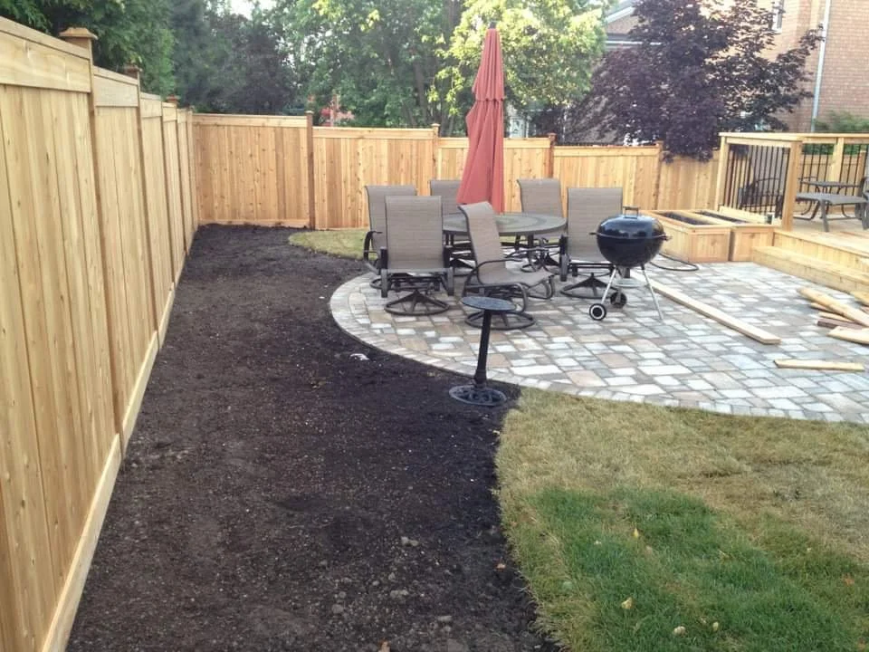 Backyard with newly installed wooden fence, patio area with paver stones, outdoor dining table with chairs, a red umbrella, a black charcoal grill, and a fire pit, with some wood and construction materials nearby.