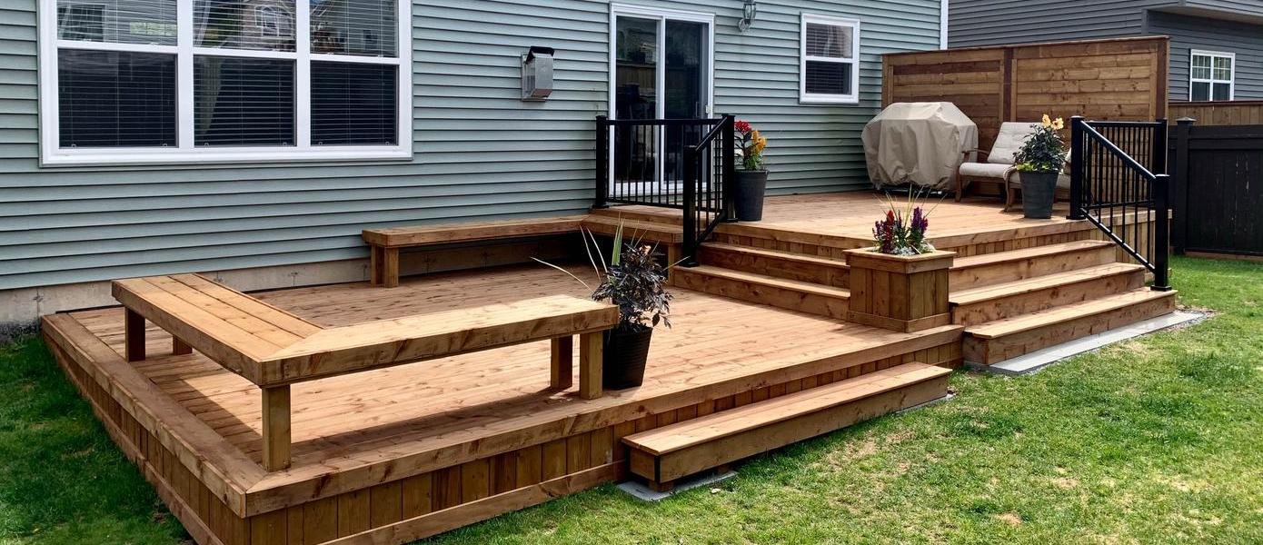Newly constructed wooden deck with multiple levels, stairs, benches, potted plants, and a covered grill next to a house with blue siding.