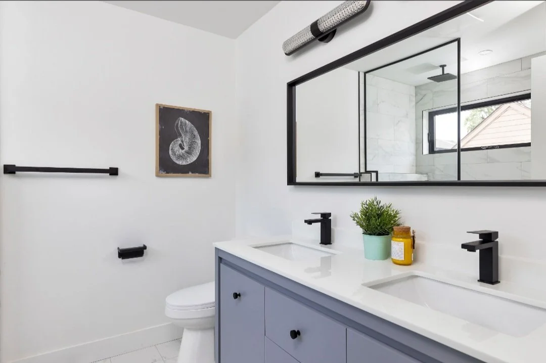 Modern bathroom with dual sinks, black fixtures, a large rectangular mirror, a small potted plant, and a walk-in shower with a window and glass enclosure.