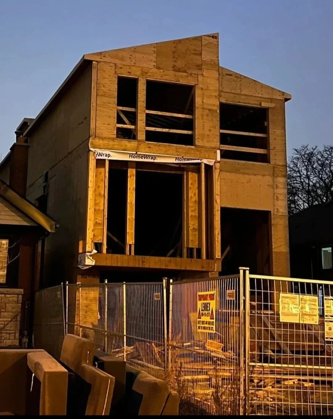 A house under construction with wooden framing in progress, surrounded by a metal fence.