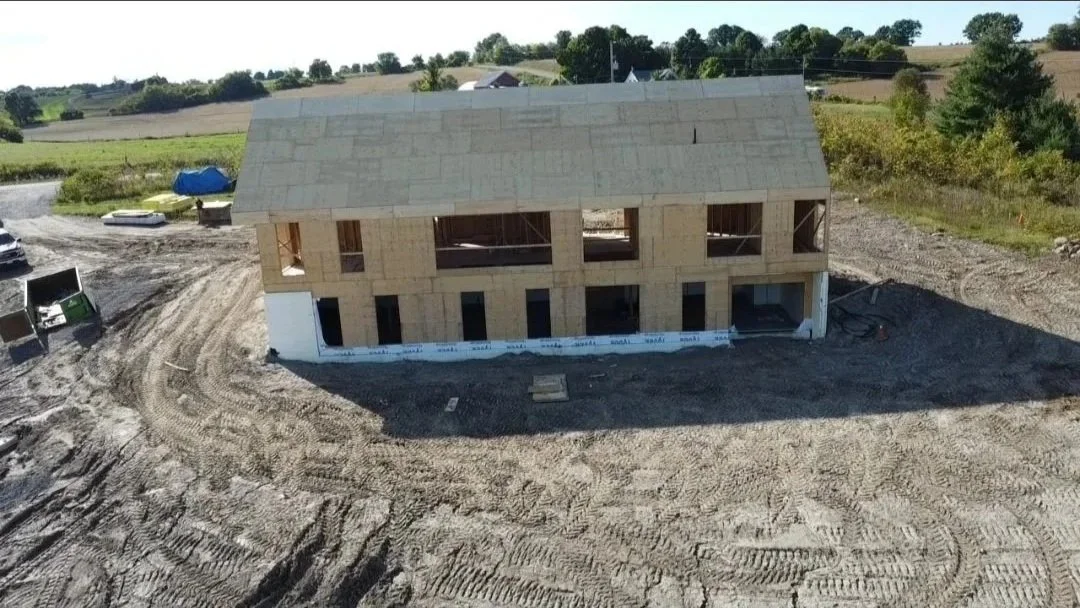 A two-story house under construction with unfinished walls and roof, surrounded by dirt and construction equipment.
