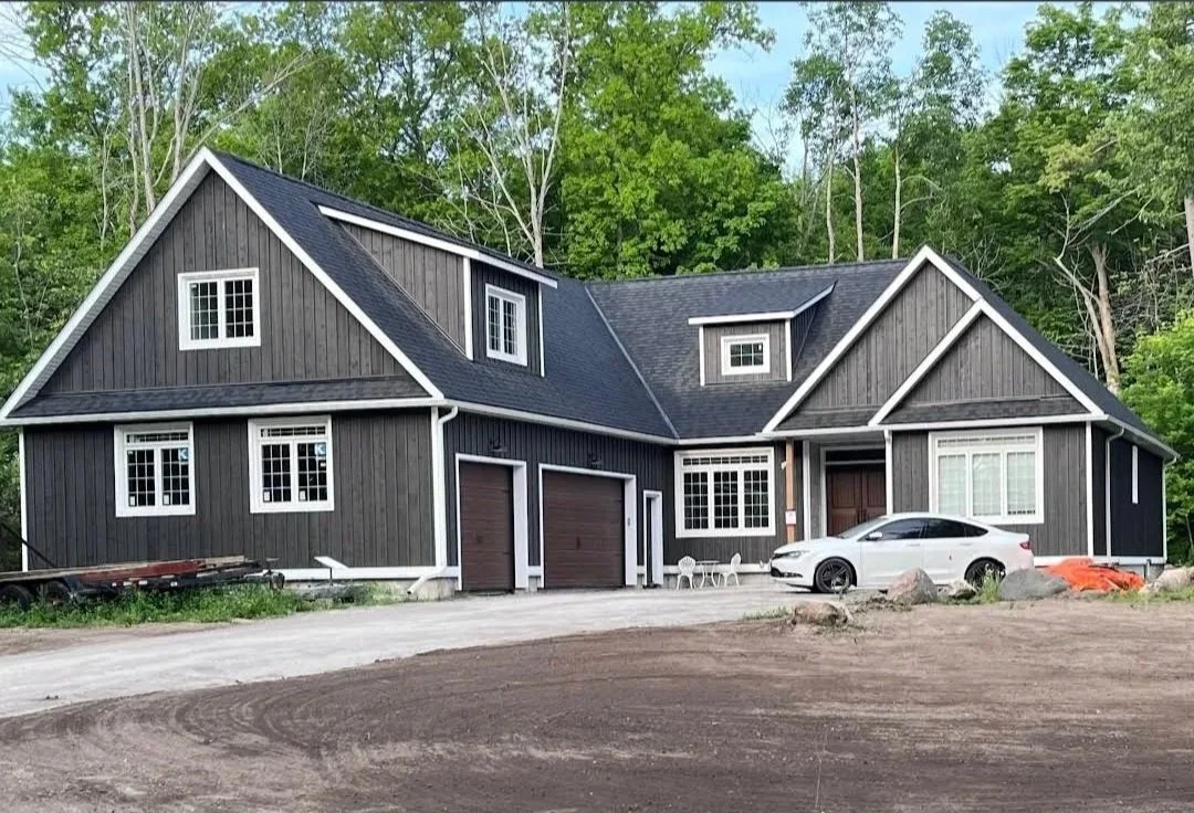 Black two-story house with white trim, large garage doors, and a white car parked in front, surrounded by trees and dirt driveway.