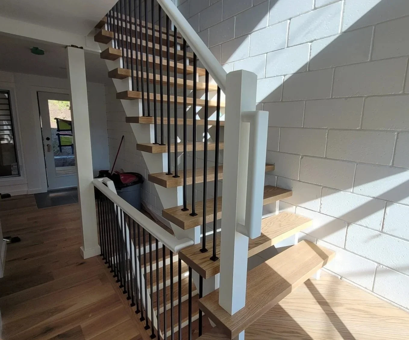Interior view of a staircase with wooden steps, white sides, black metal railings, and a white wall with brick pattern, sunlight casting shadows on the wall.