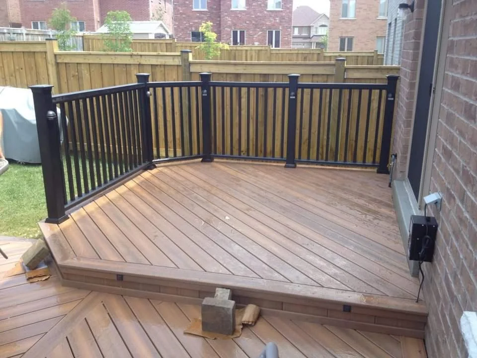 Newly built wooden deck with black metal railing attached to a brick house. There are some bricks and tools nearby, and a fenced backyard is visible in the background.