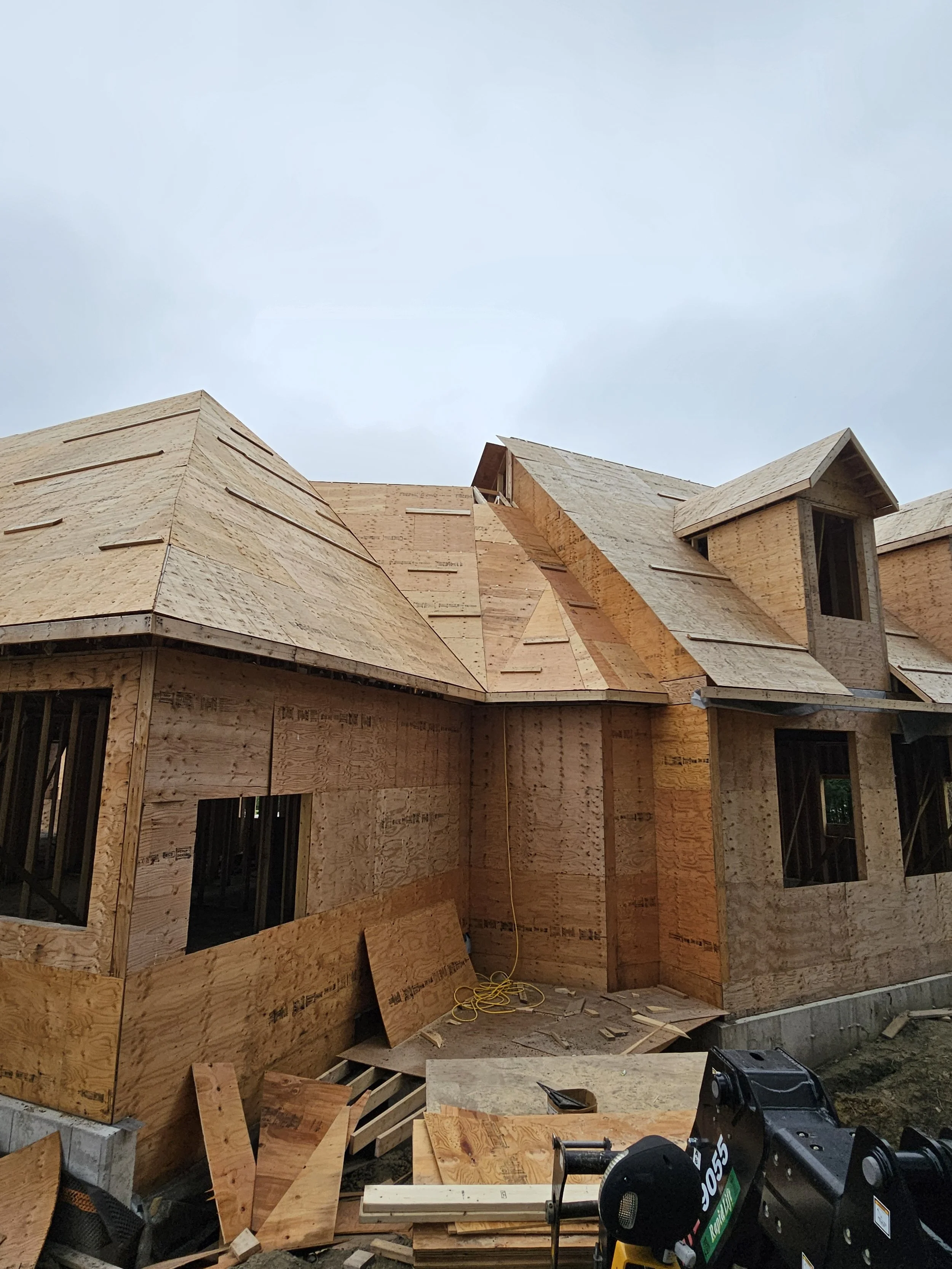 House under construction with plywood sheathing and incomplete roof, construction tools and materials on site.