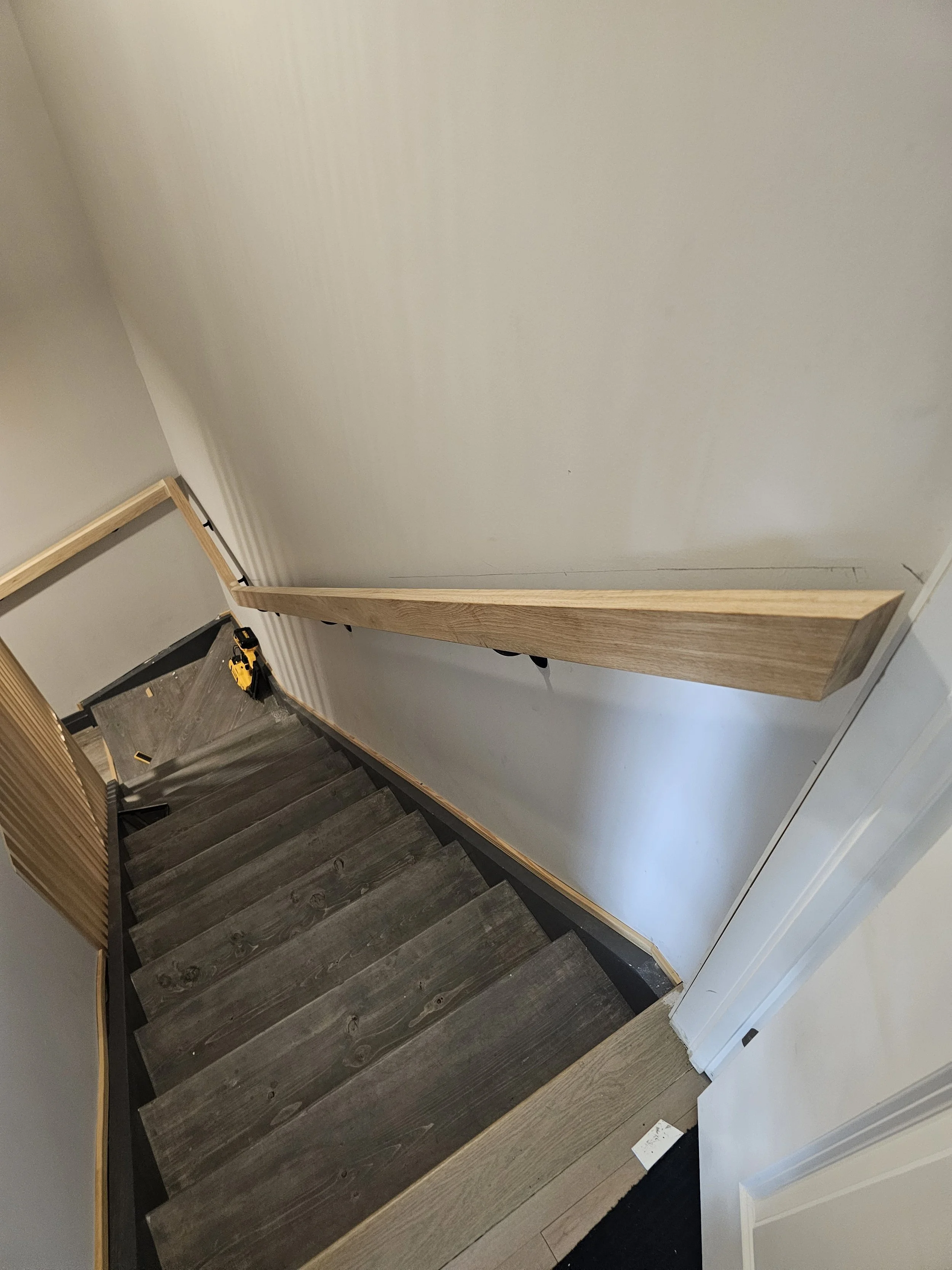View of a staircase with dark wood steps, a handrail on the right, and a cordless drill placed on the floor near the top of the stairs.