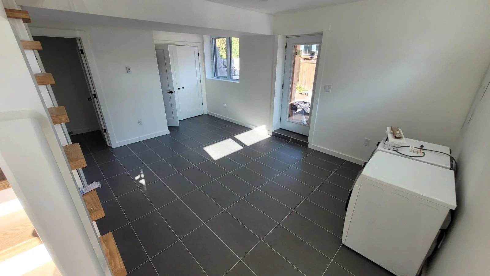 Empty laundry room with white walls, black tiled floor, a white washing machine on the right, a window on the back wall, and a door leading outside.