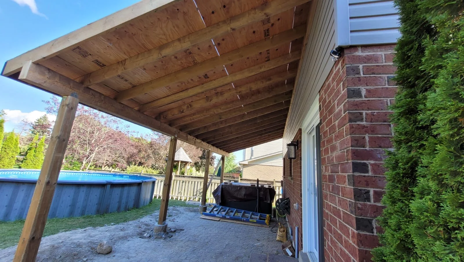 Backyard patio with a wooden porch ceiling under construction, brick house wall, and a fenced yard with a pool and trees.
