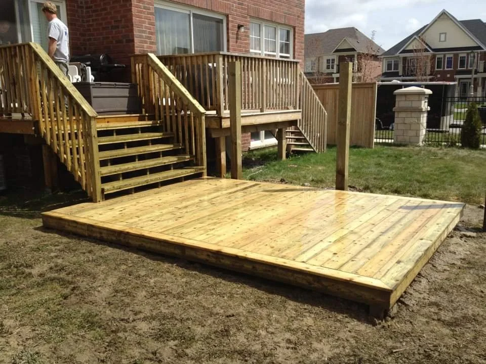A newly built wooden deck with stairs attached to a brick house, surrounded by a grassy yard, in a residential neighborhood.