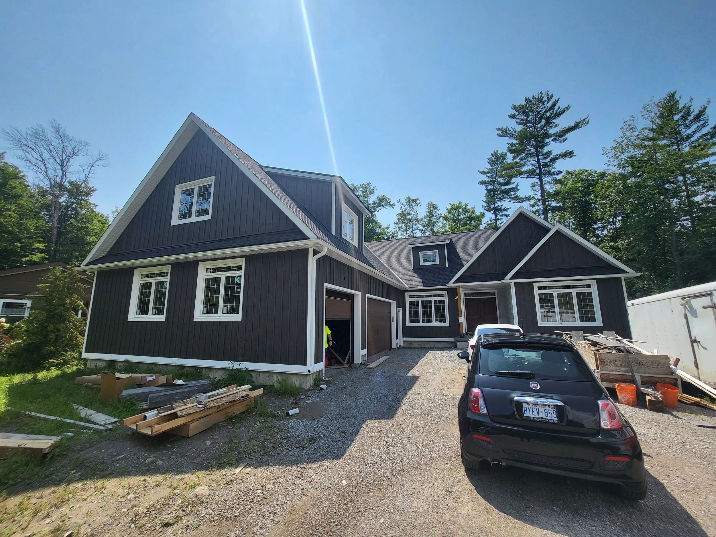 A new black house with white trim is under construction with a gravel driveway, multiple windows, a garage, and parked cars, surrounded by trees and construction materials.