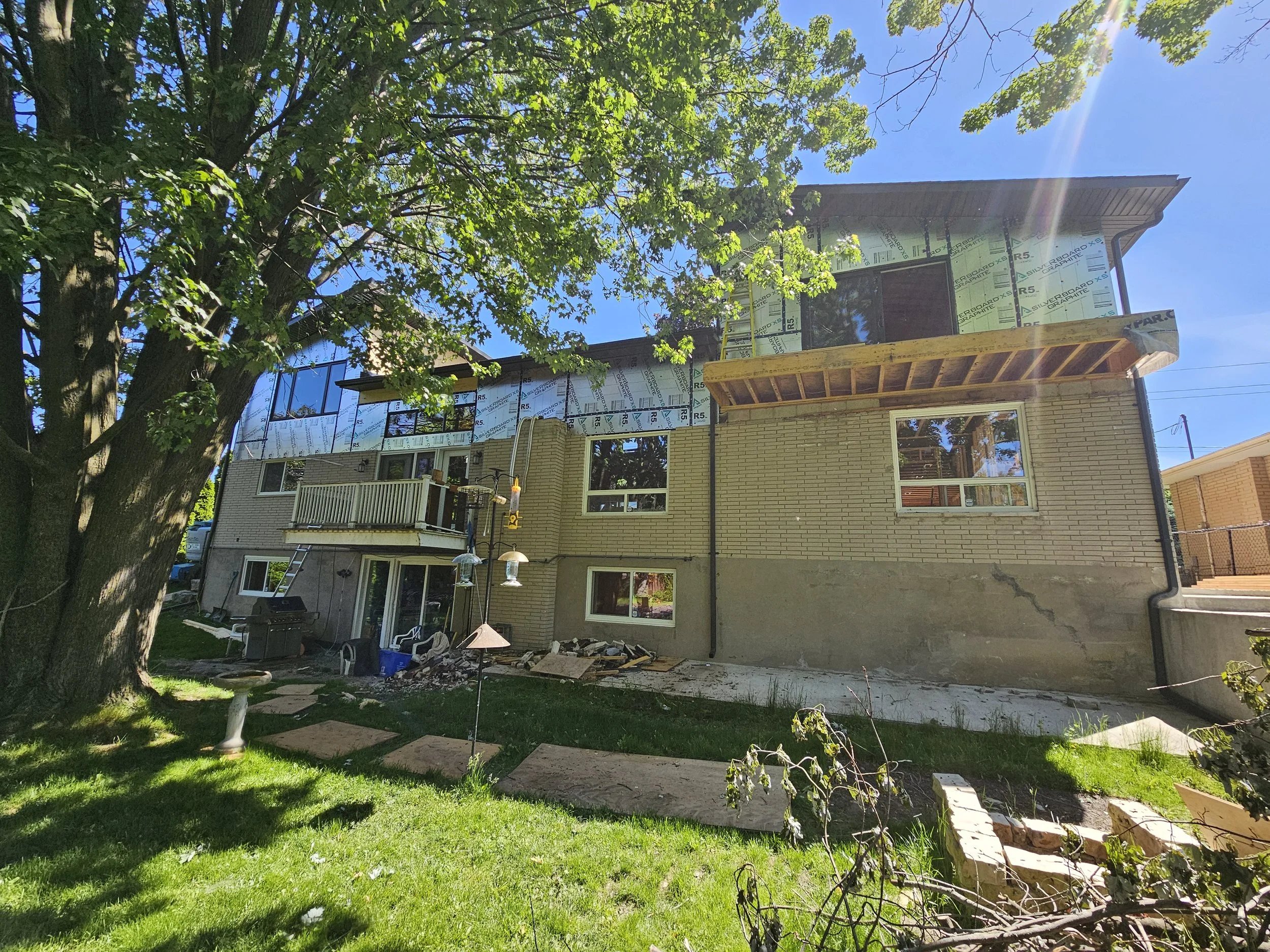 Rear view of a two-story house under construction, with exposed insulation and framing on the upper level, surrounded by a lush green yard with a large tree, bird feeders, and construction debris.