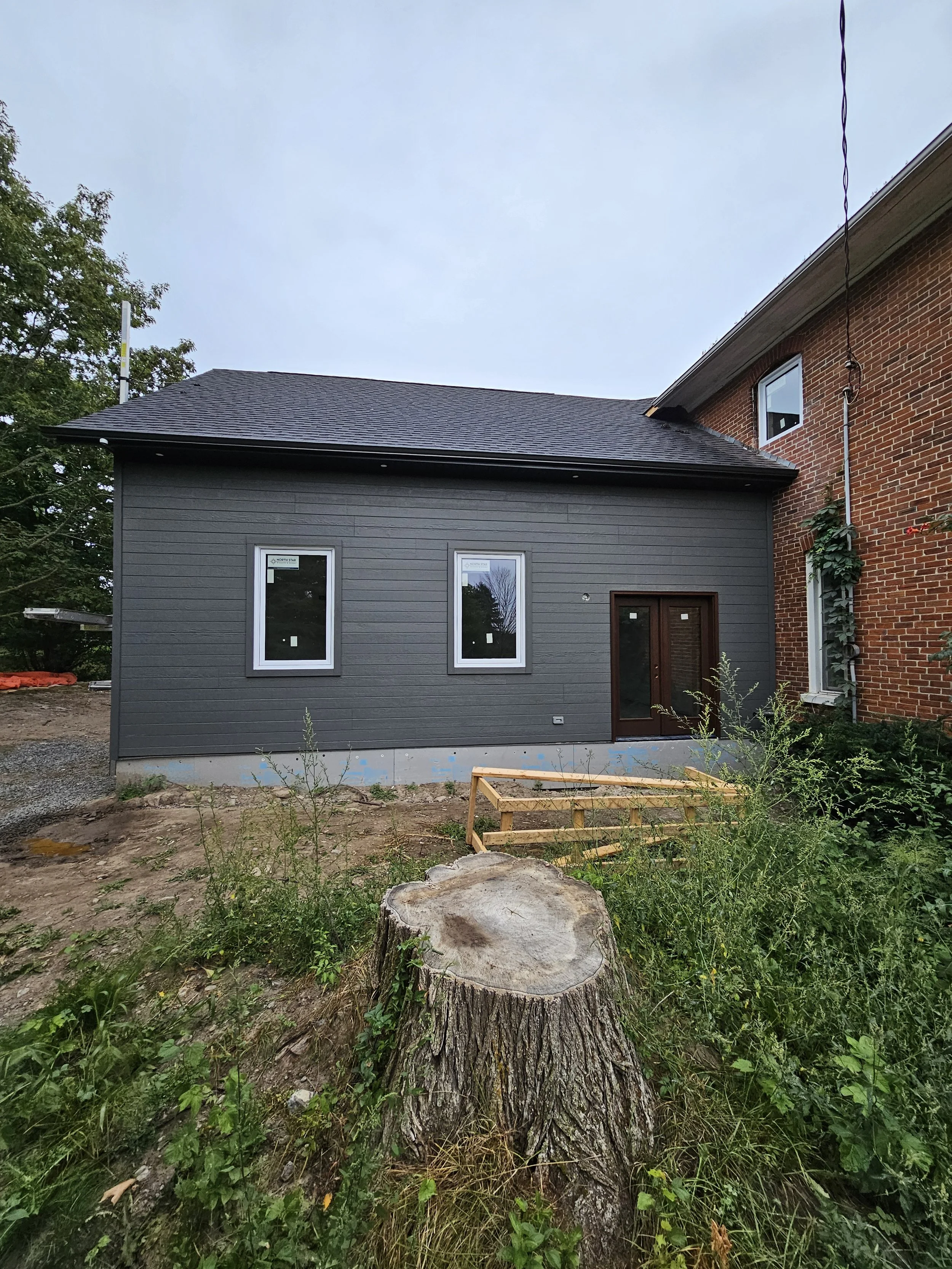 Newly constructed house with gray siding, two windows, and a wooden door, in a backyard with a tree stump and overgrown plants.