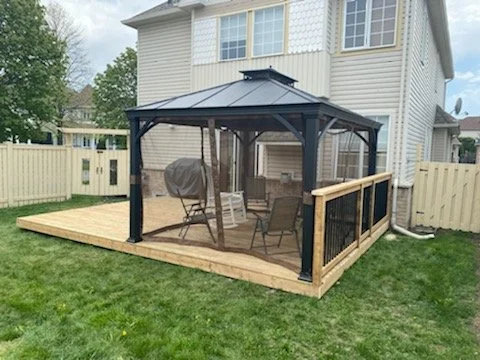 A backyard enclosed patio with a wooden deck, black metal gazebo, patio furniture, and a grill, next to a two-story house.