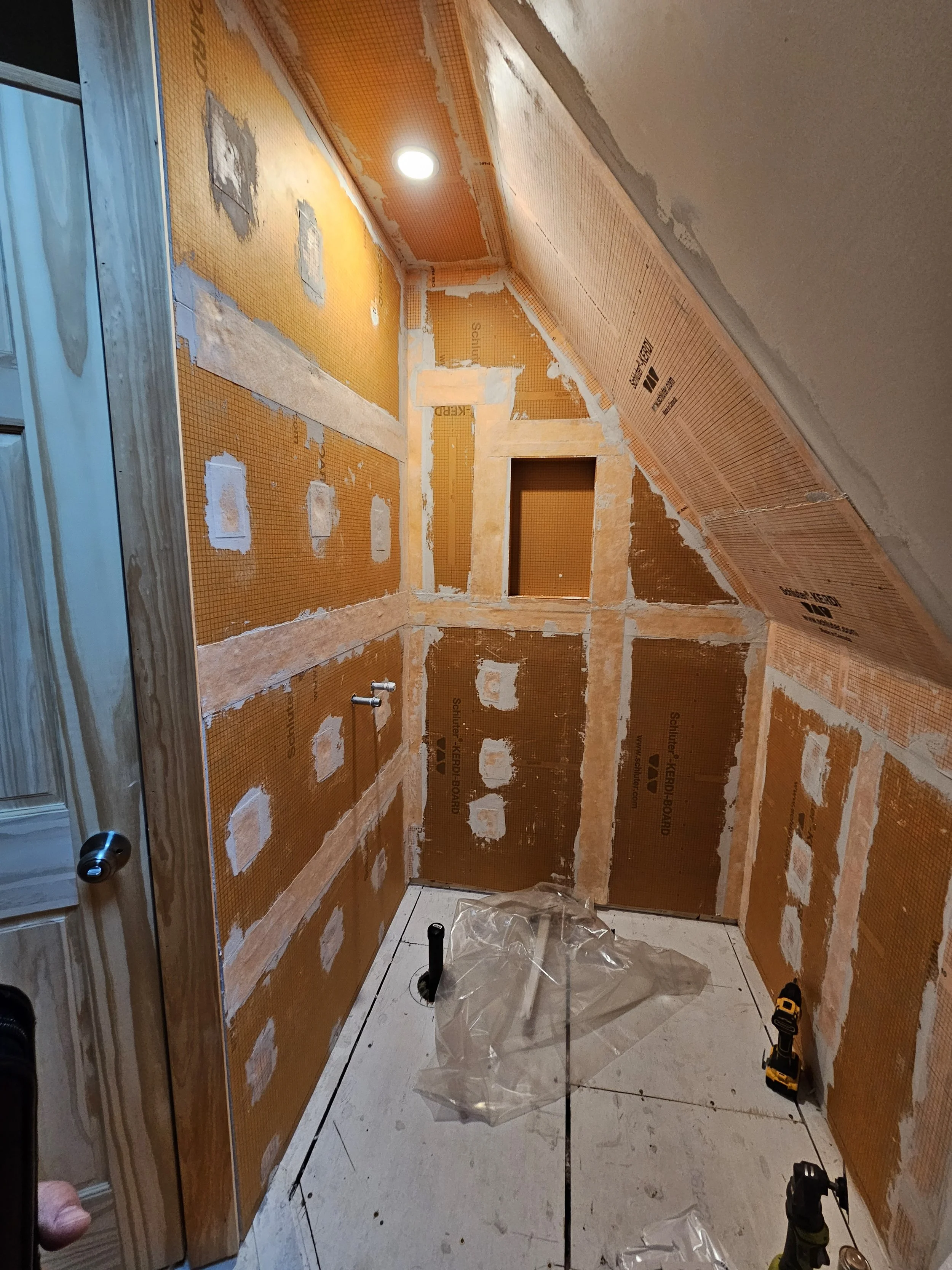 Bathroom under construction with orange drywall panels, exposed pipes, a partially installed toilet, and tools on the floor.