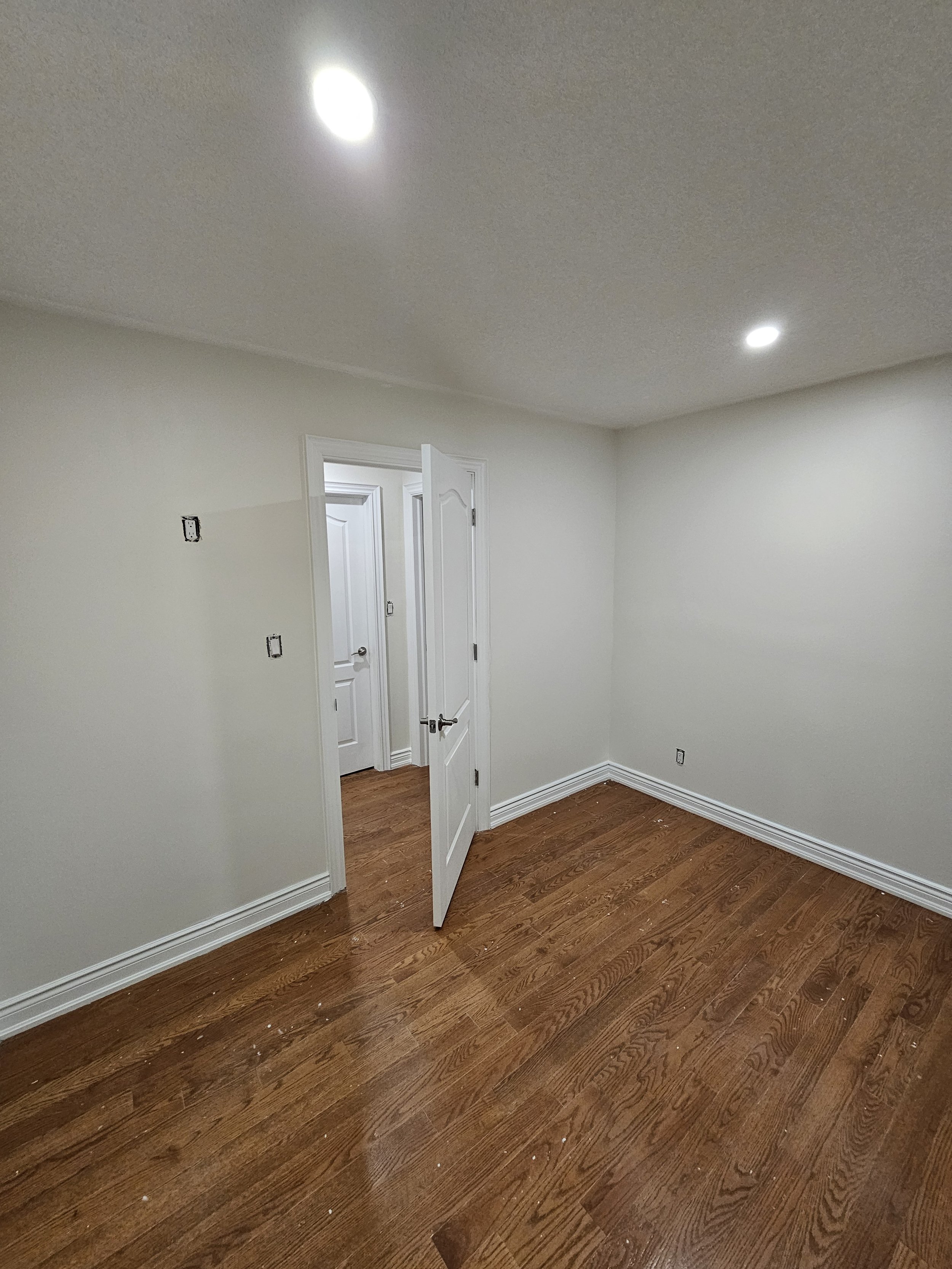 Empty room with white walls, hardwood floors, and an open door leading to a hallway with similar walls and flooring.