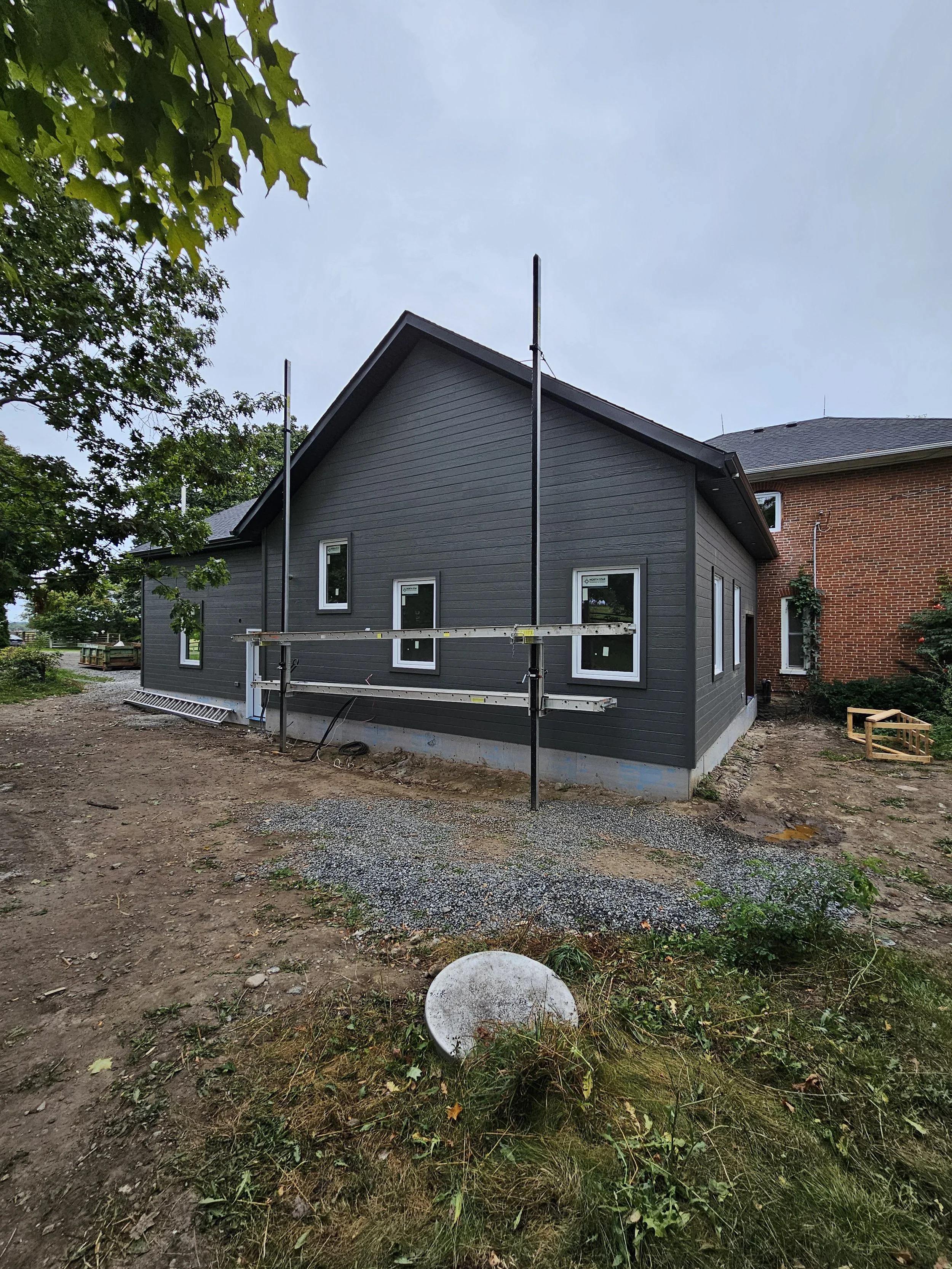 Newly constructed gray house with black trim, multiple windows, and a sloped roof, surrounded by a partially landscaped yard with scaffolding and building materials present.