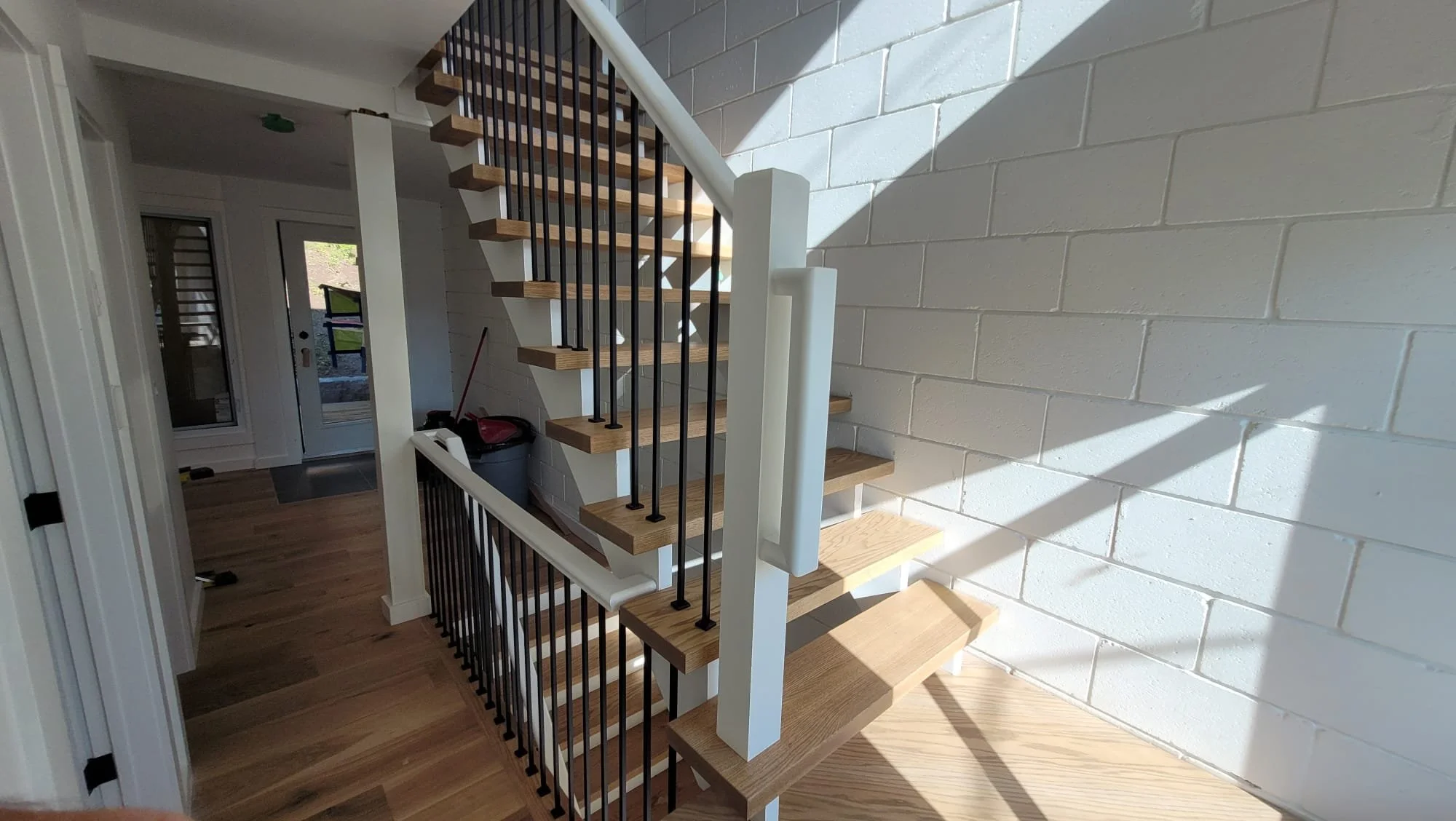 Interior view of a modern staircase with wooden steps, black metal balusters, and white handrails next to a white brick wall, with natural light casting shadows.