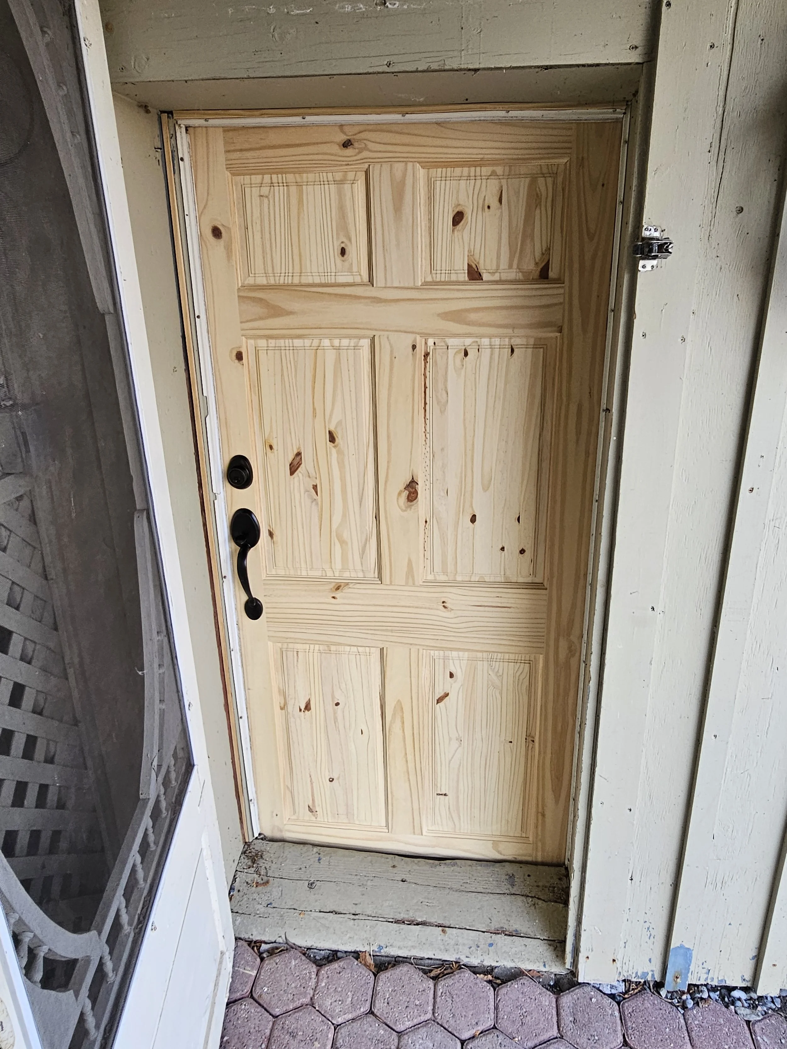 Unfinished wooden door with black handle and lock on a porch with hexagonal pavers, side wall, and a screen door on the left.