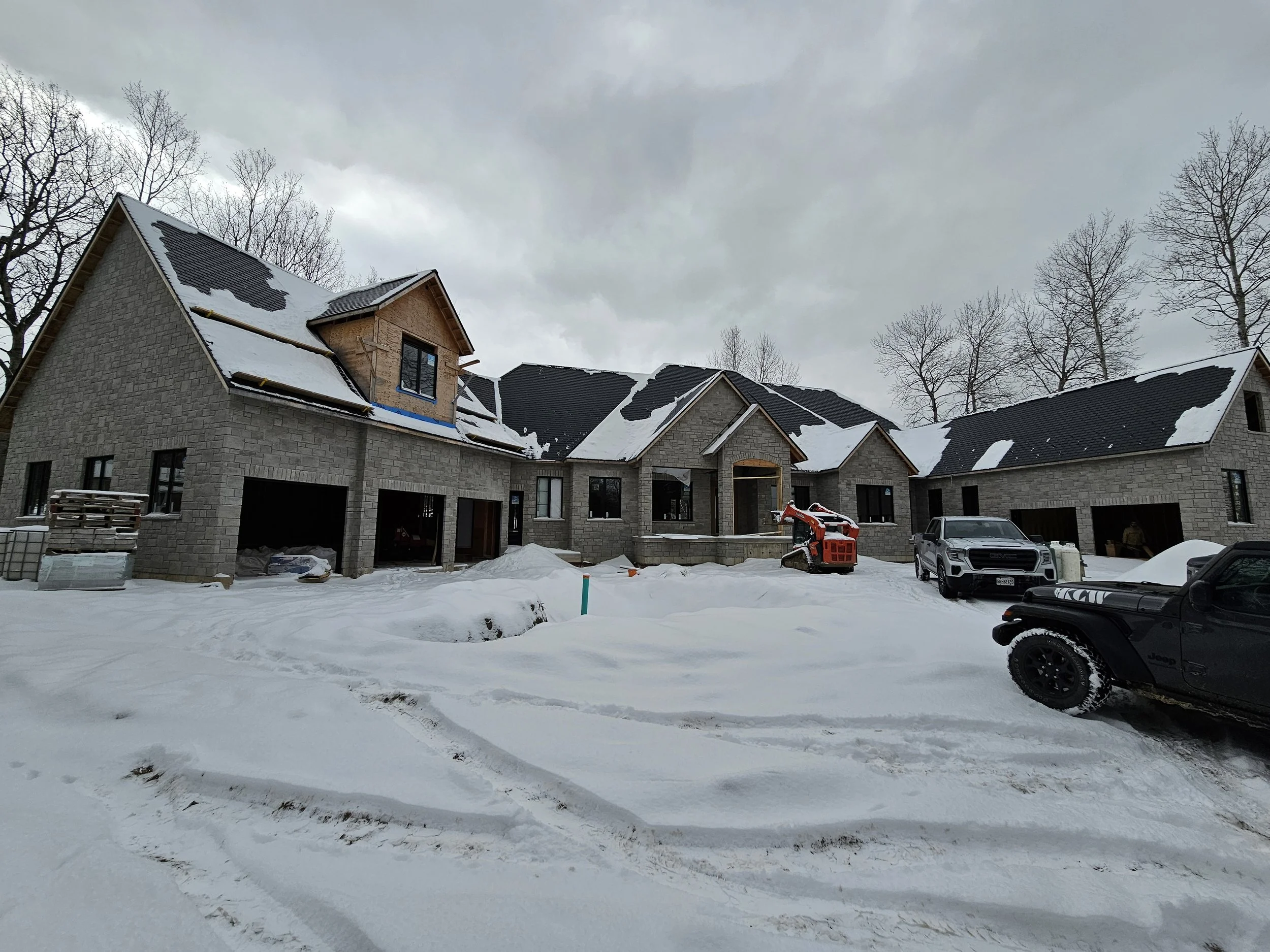 A large house under construction with a snow-covered yard, parked trucks and construction equipment, and trees without leaves in the background, all set on a cloudy winter day.