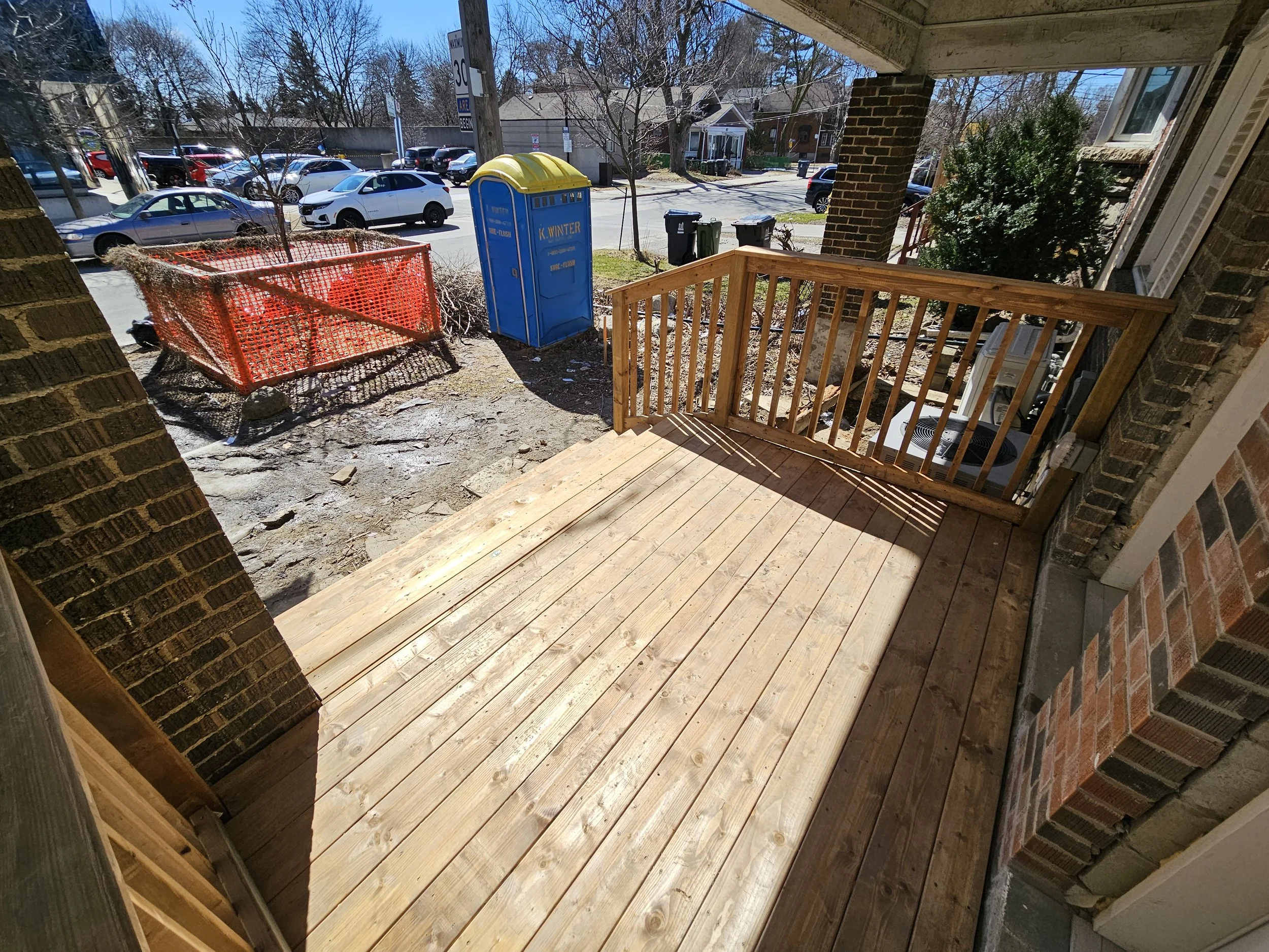 View of a new wooden porch with railing, construction materials, and construction zone outside of a brick building, with parked cars, trash bins, and a blue portable toilet in the background.