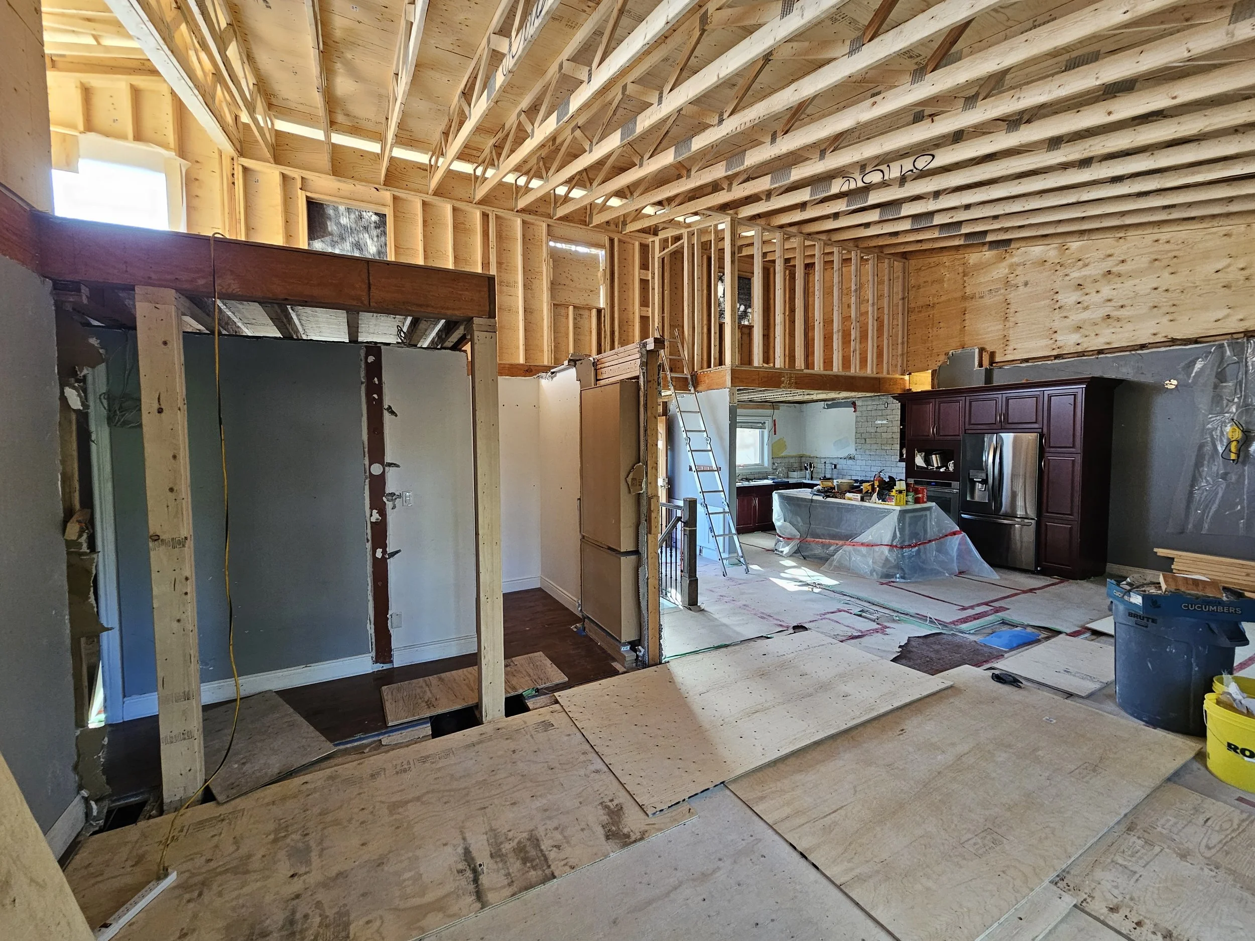 Interior view of a house under construction, showing wooden framing, an unfinished kitchen with cabinets, a ladder, and construction materials.