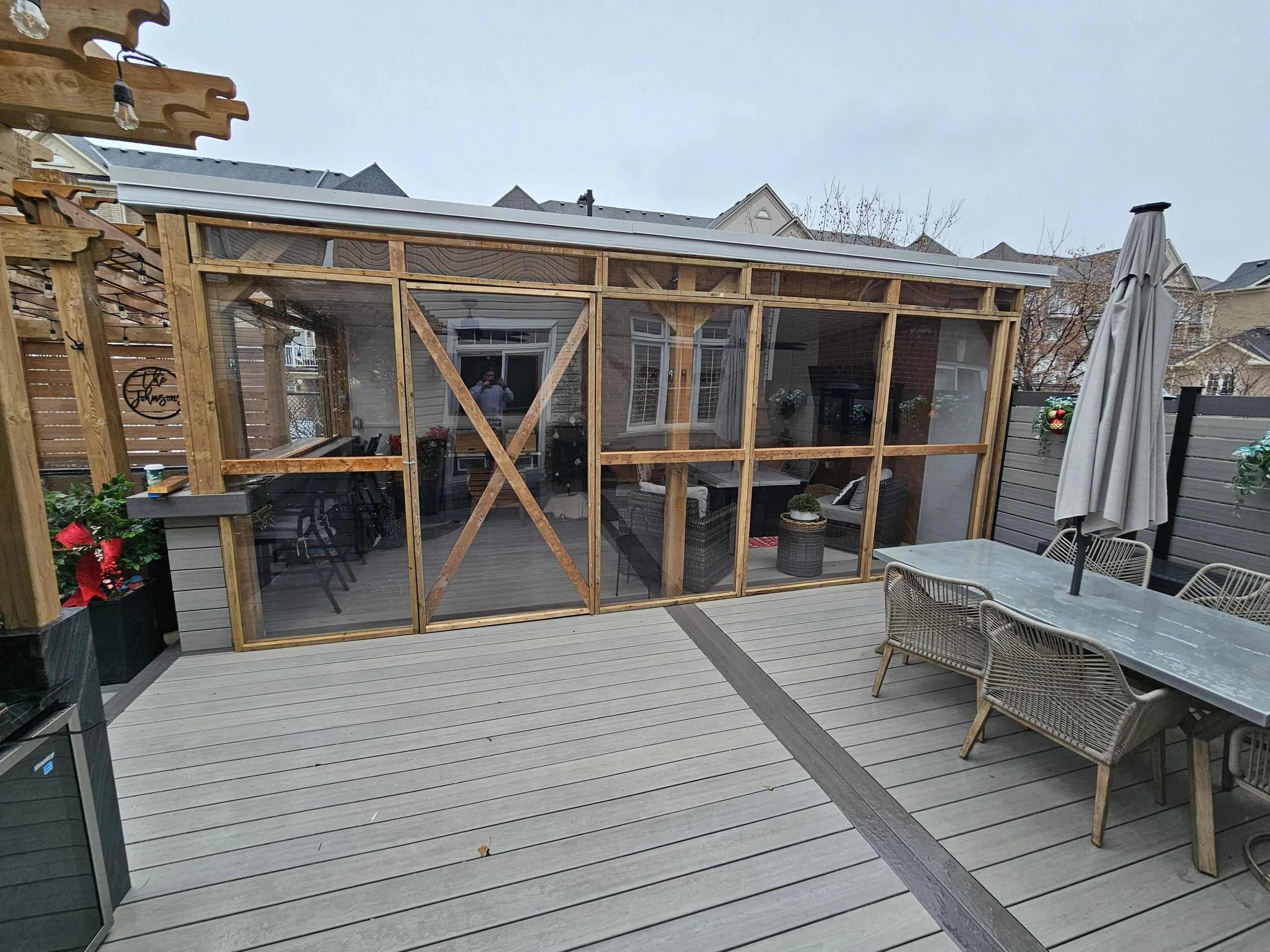 An unfinished backyard patio with a wooden and glass enclosed structure, outdoor dining table with chairs, and holiday decorations on the fence.