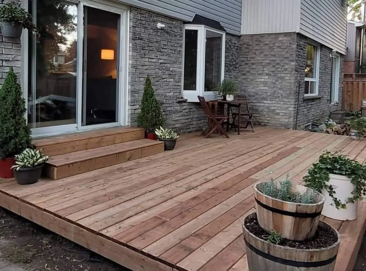 A wooden deck outside a house with sliding glass door, potted plants, a small table with two chairs, and other plants in pots.