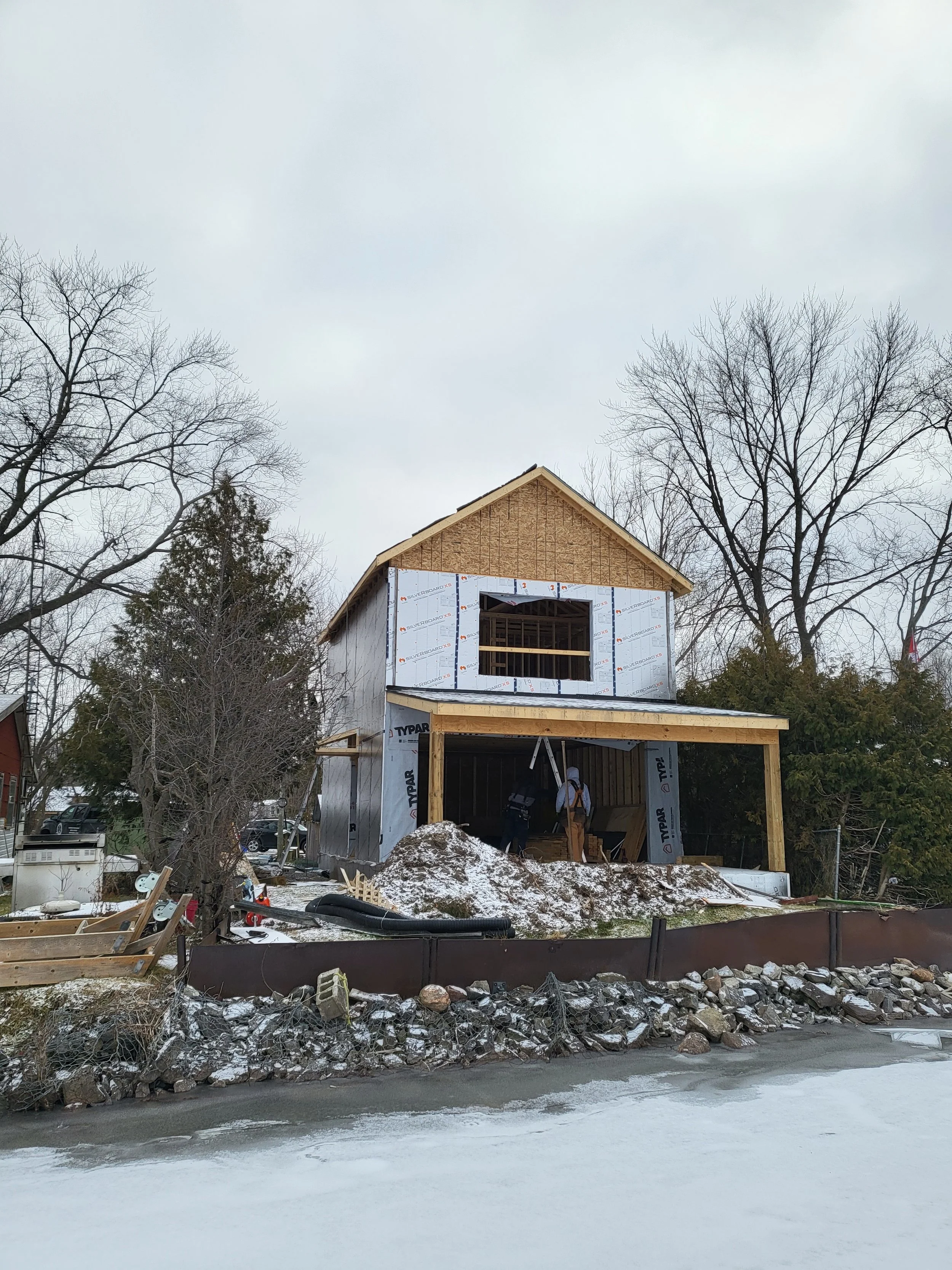 A house under construction with two stories, wooden framing, and snow on the ground, trees in the background, and workers inside the structure.
