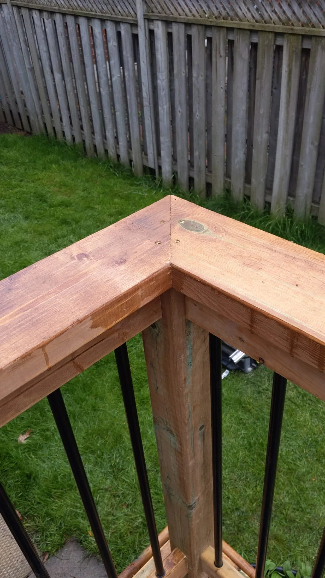 Close-up view of a wooden deck railing corner with black metal balusters, overlooking a grassy backyard with a wooden fence.
