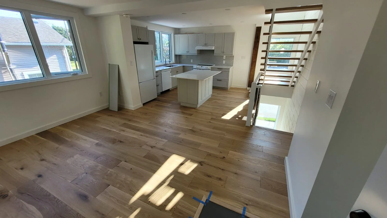 Interior view of a modern, spacious kitchen with light-colored cabinets, a small island, large windows, and hardwood floors. A staircase with wooden steps and white risers is visible to the right.