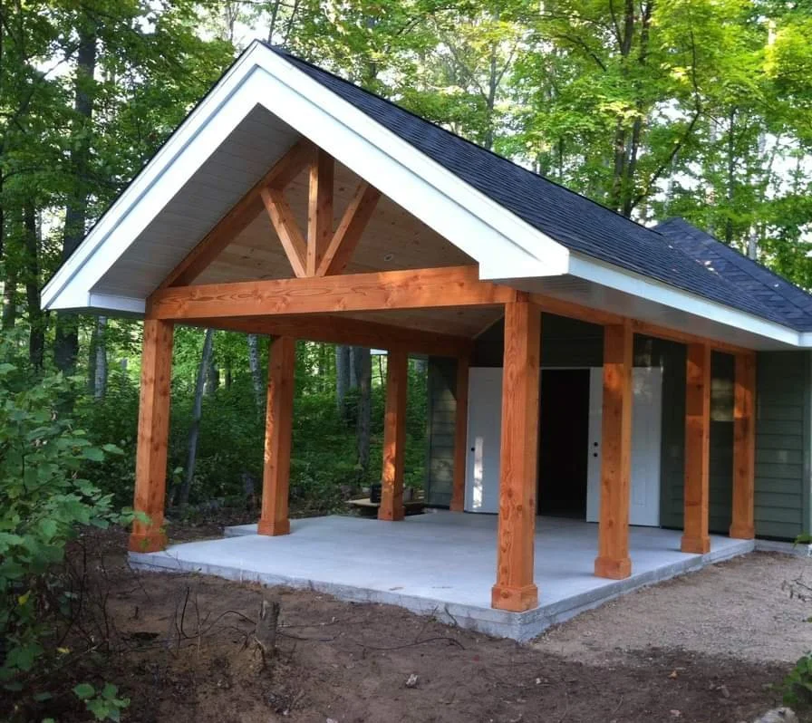 A house under construction with a wooden porch supported by large timber posts, a concrete slab foundation, and a black shingled roof, surrounded by green trees.