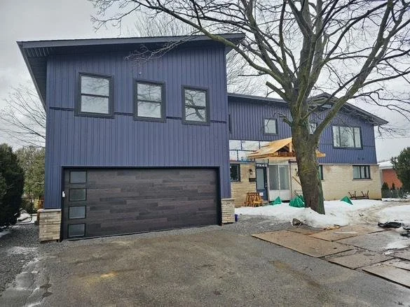 Two-story house with black exterior and a large garage door, surrounded by snow and a large tree in the yard.