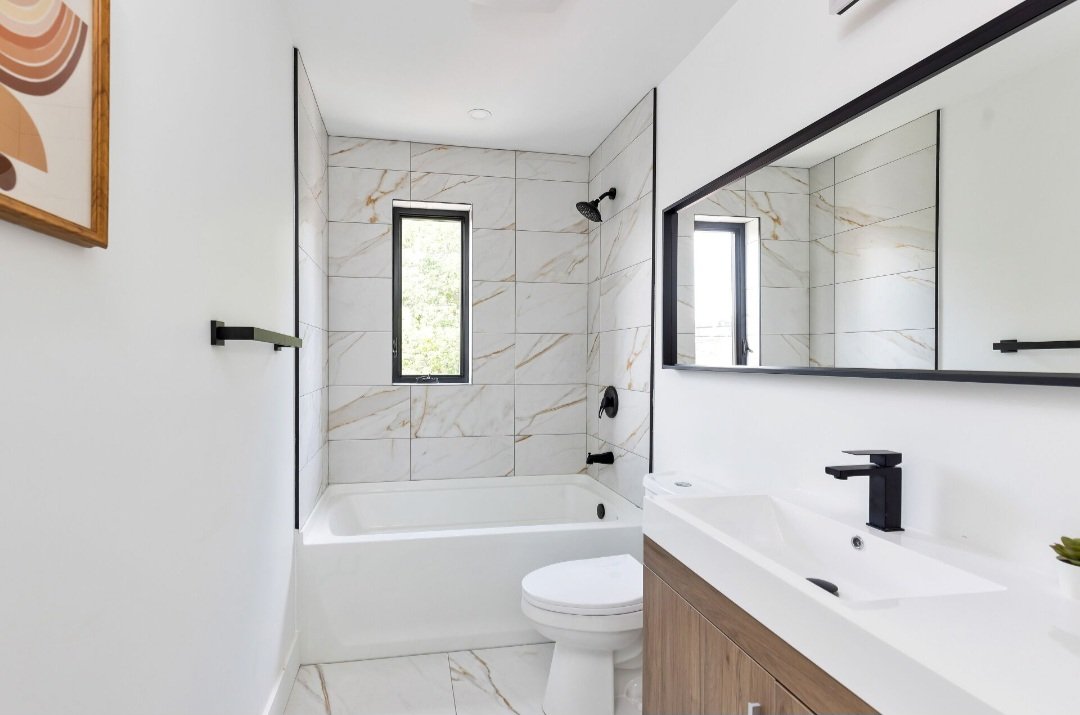 Modern bathroom with a bathtub, white walls, marble tiles, a small vertical window, a black showerhead, a large mirror, a white sink with a black faucet, and a wooden cabinet.