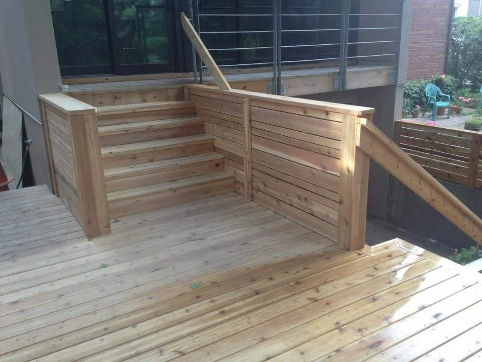 Wooden stairs leading to a raised deck with a railing, connecting to a house with a glass door and a balcony.